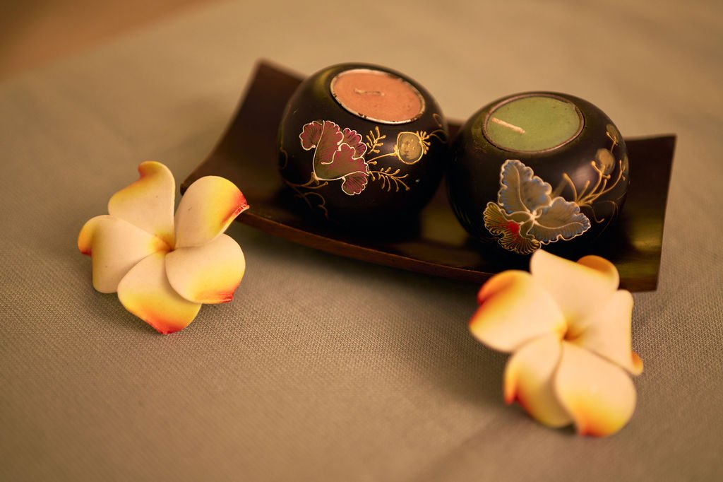 Deux bougies colorées avec des motifs floraux, placées sur une petite barque, accompagnées de deux fleurs blanches avec des extrémités rougeâtres.