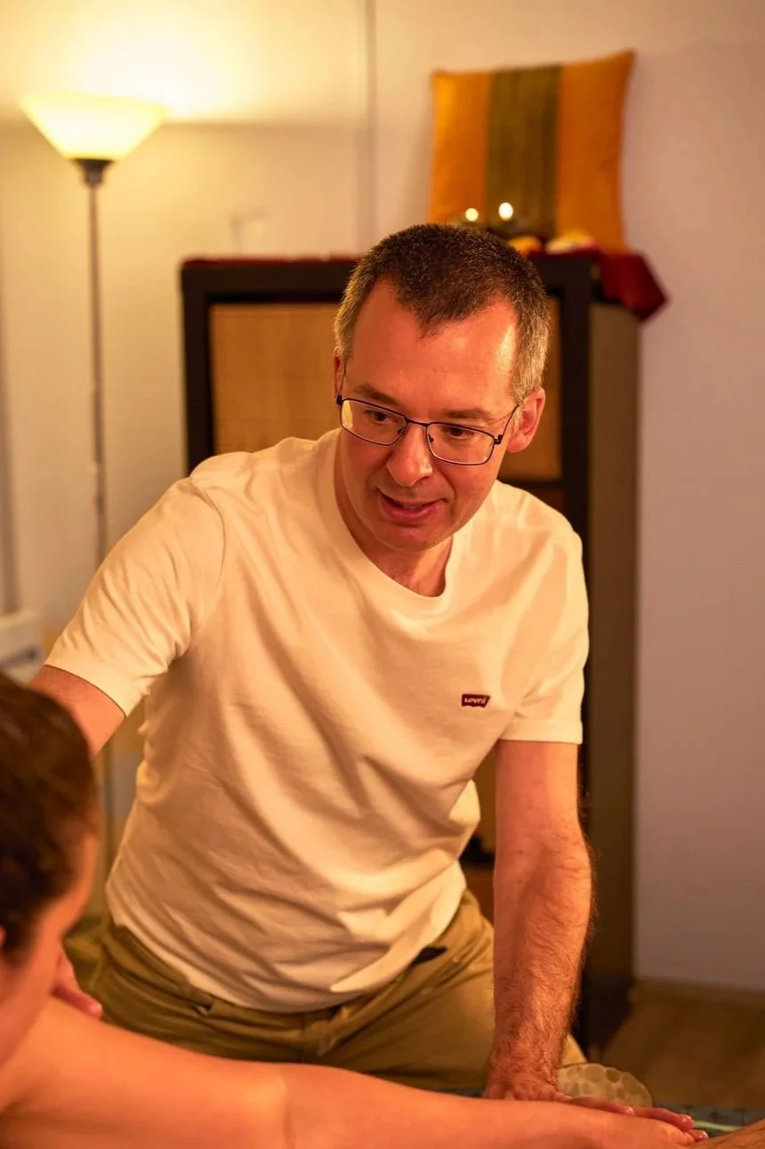 Un homme portant des lunettes et un t-shirt blanc est assis, engageant une conversation ou montrant quelque chose à une autre personne. Il se trouve dans une pièce chaleureuse avec une lampe sur pied et un panneau décoratif en arrière-plan.