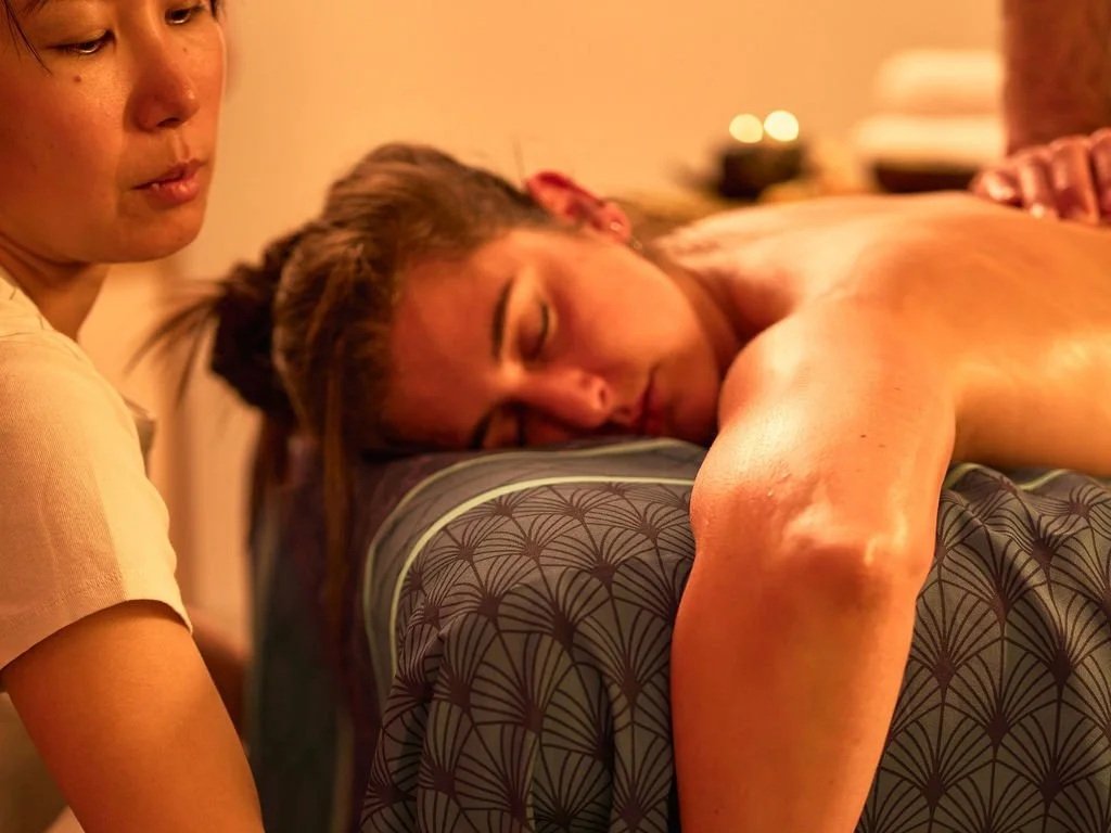 Une femme allongée sur une table de massage avec un visage détendu, pendant qu'une autre femme, probablement une masseuse, se tient à côté d'elle dans un environnement chaleureux et relaxant.