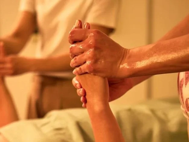 Deux mains se serrent, une semble en massage ou en soin, dans un environnement chaud et détendu.