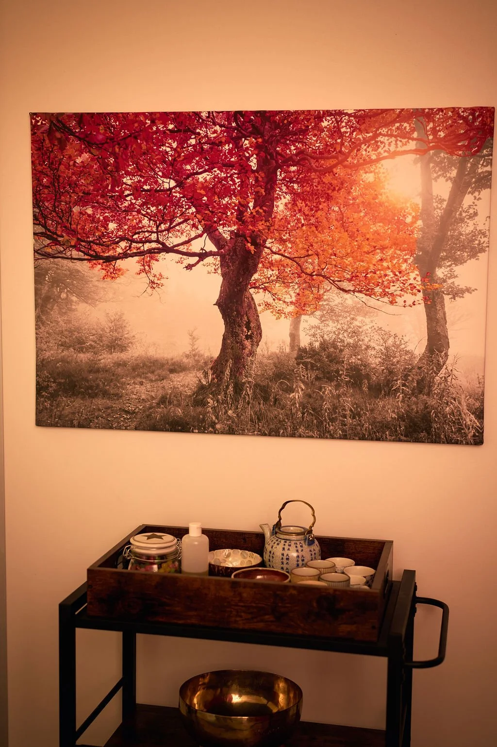 Une peinture représentant un arbre aux feuilles rouges dans une forêt brumeuse avec le soleil levant. En dessous, une table en bois avec des objets décoratifs, notamment une théière, des tasses et un flacon.
