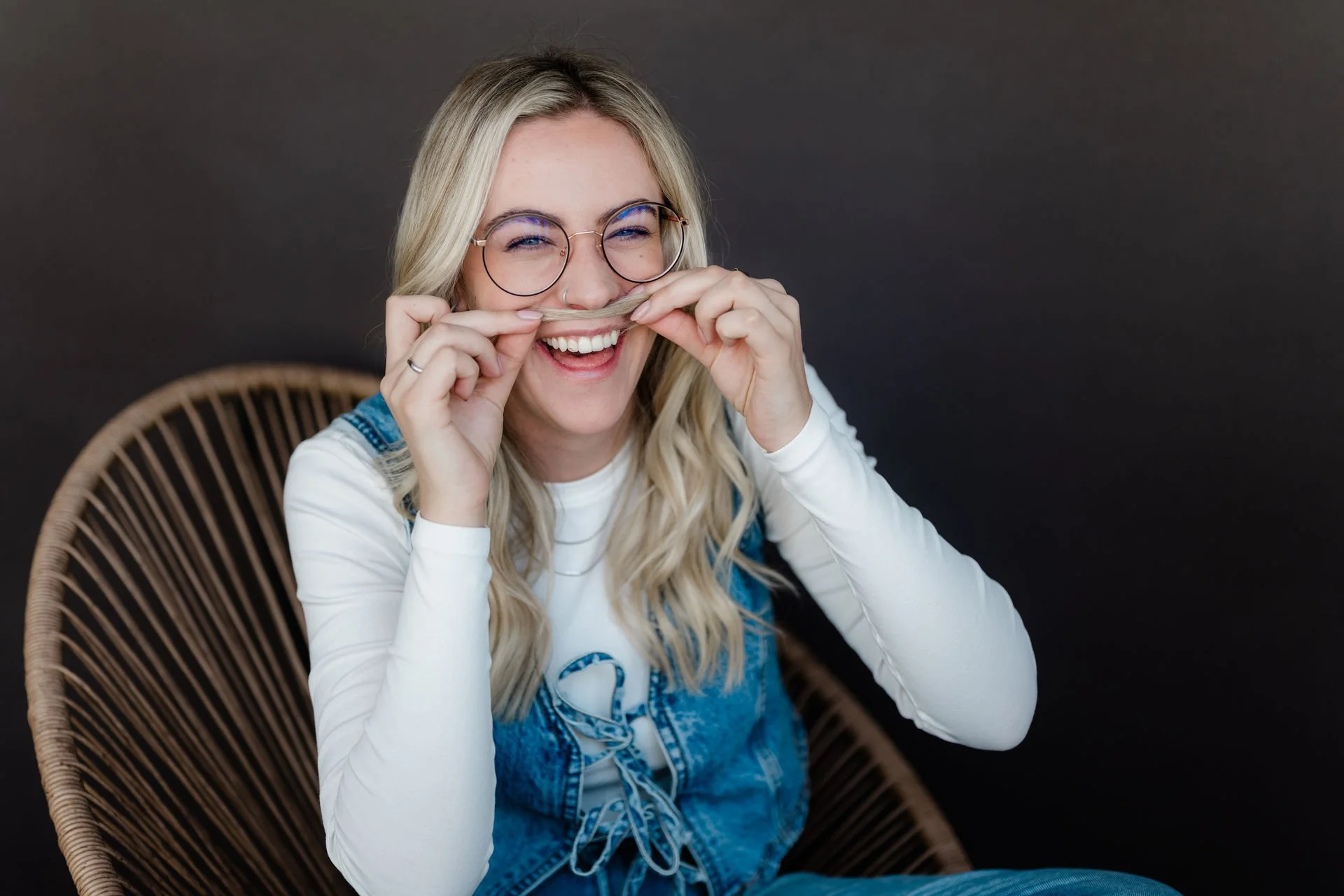 Lächelnde junge Frau mit Brille, die Haare mit einem Haarbügel als Nase benutzt und lachend sitzt in einem Korbsessel vor dunklem Hintergrund.