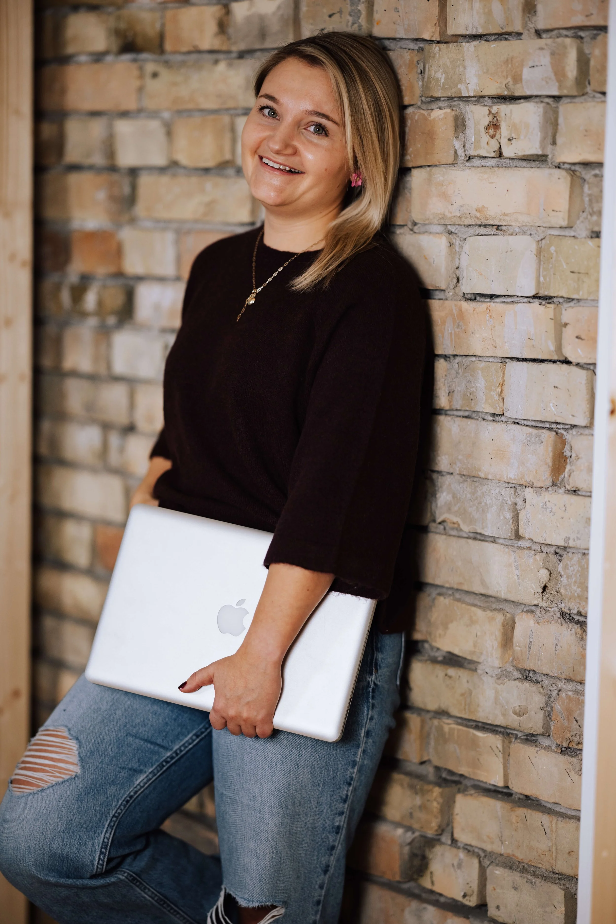Eine junge Frau lehnt lächelnd an einer Ziegelmauer, trägt eine schwarze Oberbekleidung, eine Halskette und trägt einen MacBook-Laptop in der Hand. Sie hat blondes Haar und trägt pinke Ohrringe.