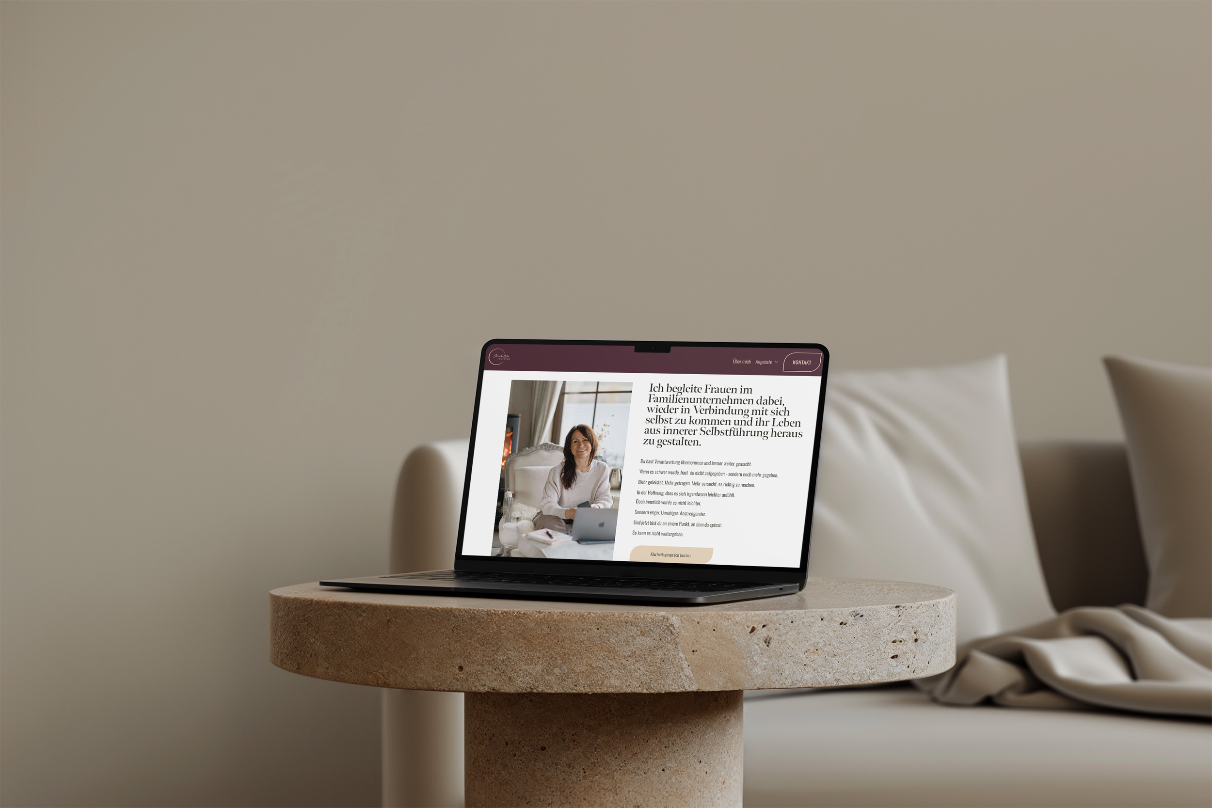 Laptop auf einem runden, grauen Stehtisch. Der Laptop zeigt eine Webseite mit einem Text in deutscher Sprache. Im Hintergrund ist eine weiße Couch und eine beige Decke sichtbar.