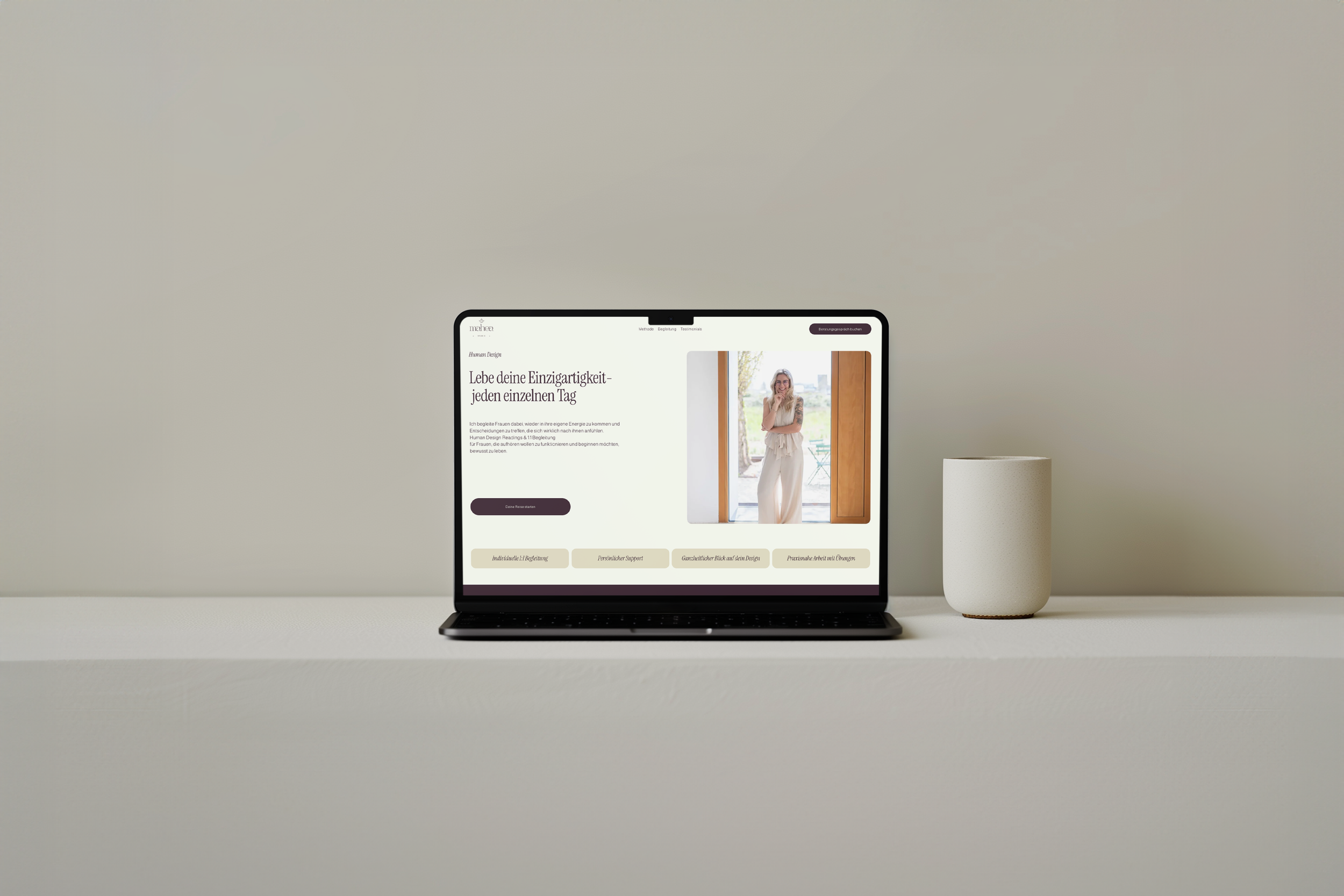 Ein Laptop auf einem weißen Schreibtisch, zeigt eine Webseite mit einer Frau, die vor einem Fenster steht. Neben dem Laptop befindet sich eine weiße Tasse.