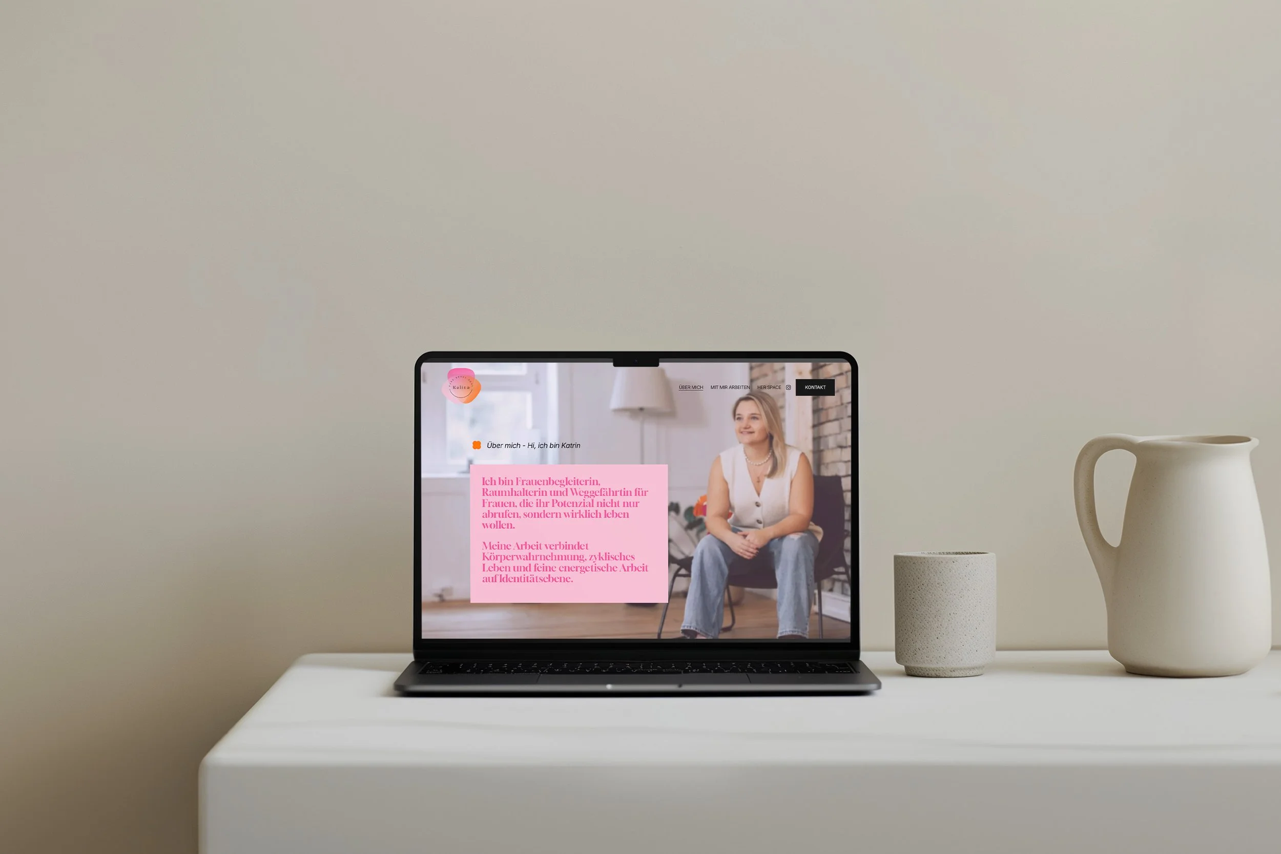 Ein Laptop auf einem weißen Tisch, der eine Webseite mit einer Frau im Gespräch zeigt, umgeben von einer Kerze und einer Vase, gegen einen cremefarbenen Hintergrund.