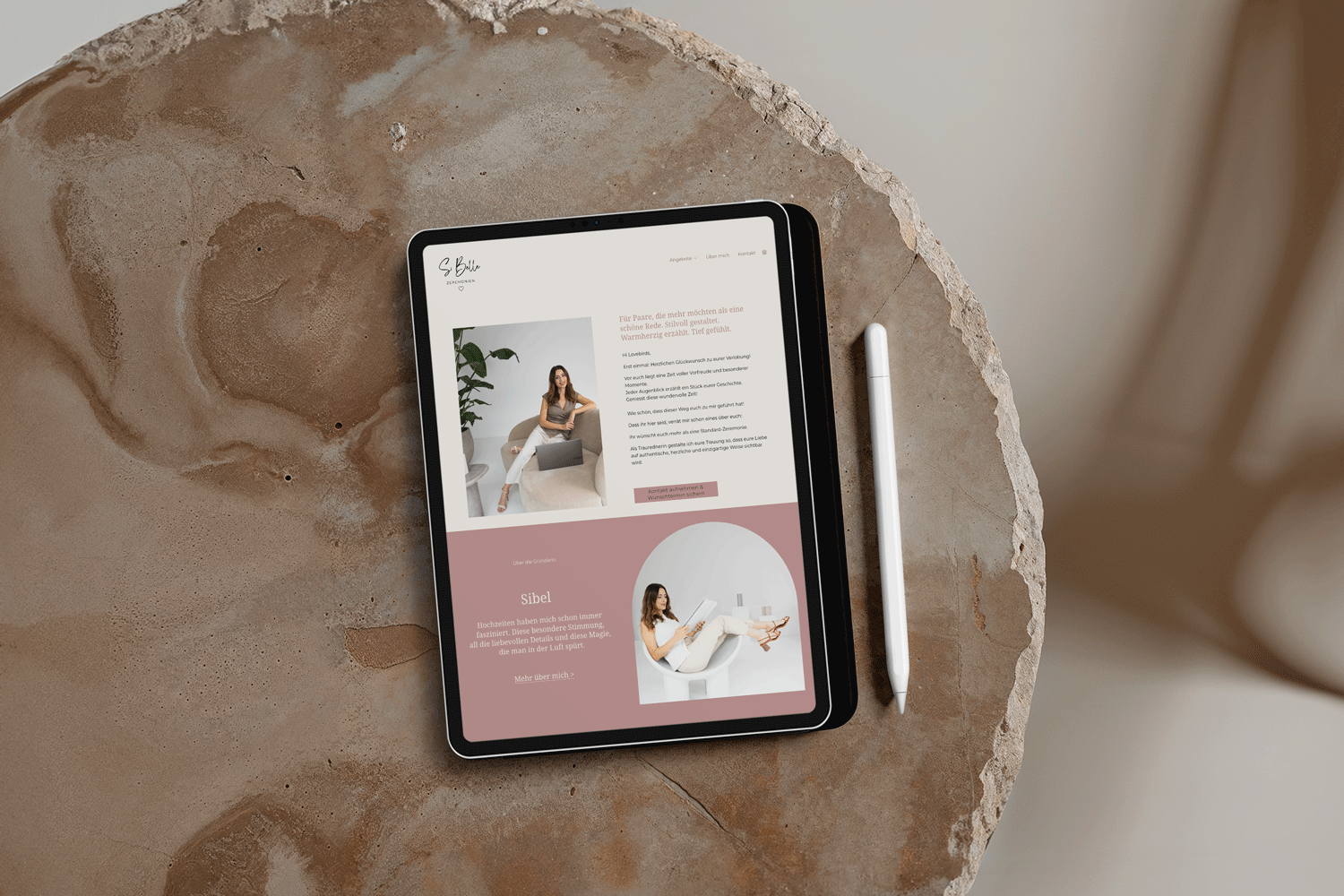 Ein Tablet mit einer Webseite über eine Frau namens Sibel, die auf einem grauen Sofa sitzt, mit einem Laptop. Das Bild ist auf einem runden Steintisch mit einem weißen Stift neben dem Tablet. Die Webseite hat ein freundliches Design in Rosa- und Erdtönen.