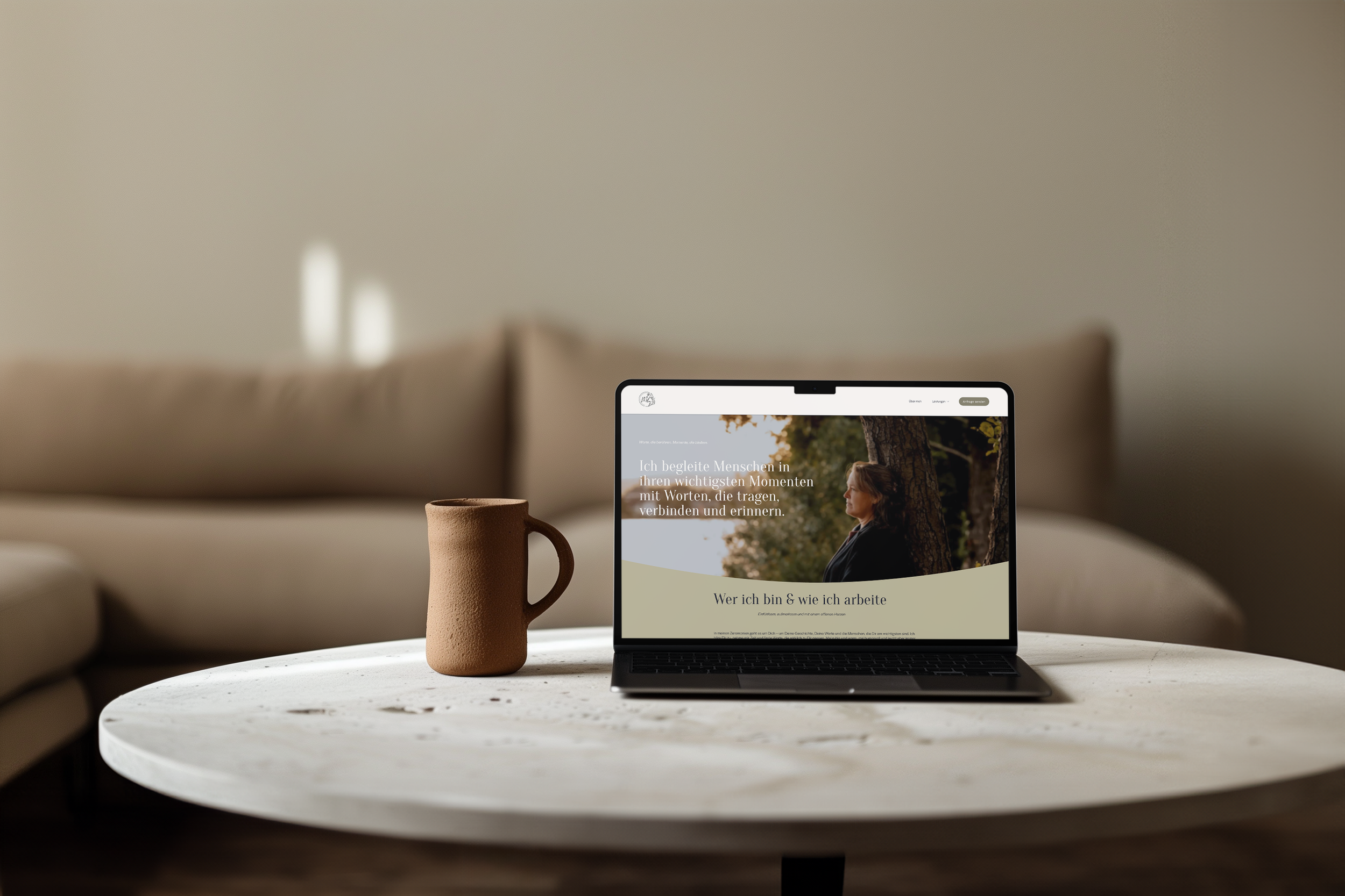 Ein Laptop zeigt eine Webseite mit einer Frau, die an einem Baum im Freien sitzt, auf einem weißen Couchtisch. Daneben steht eine braune Keramik-Tasse, im Hintergrund ein beige Couch im Wohnzimmer.