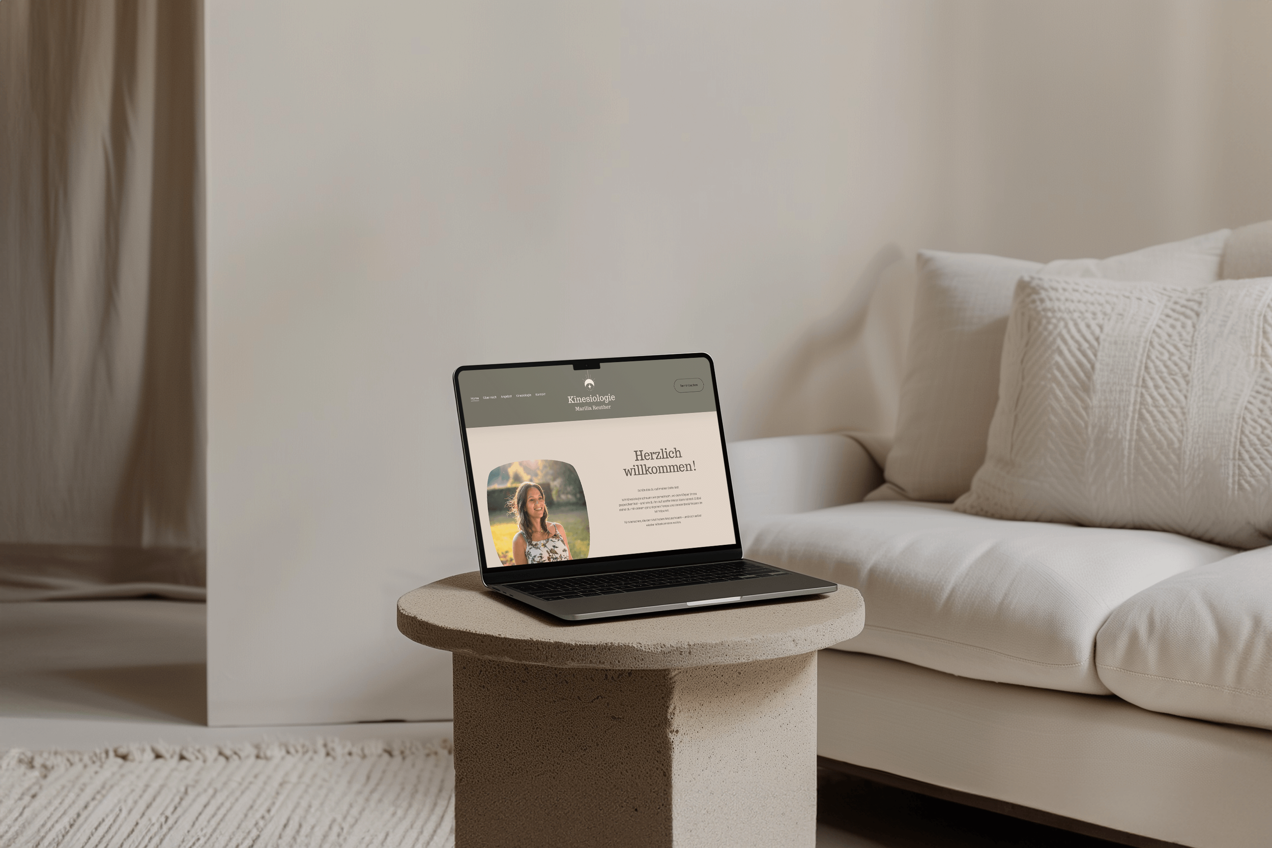 Laptop auf einem runden, beigefarbenen Tisch in einem hellen Wohnzimmer, auf dem Bildschirm ist eine Webseite mit einem Foto einer Frau und dem Text 'Herzlich willkommen!' zu sehen.