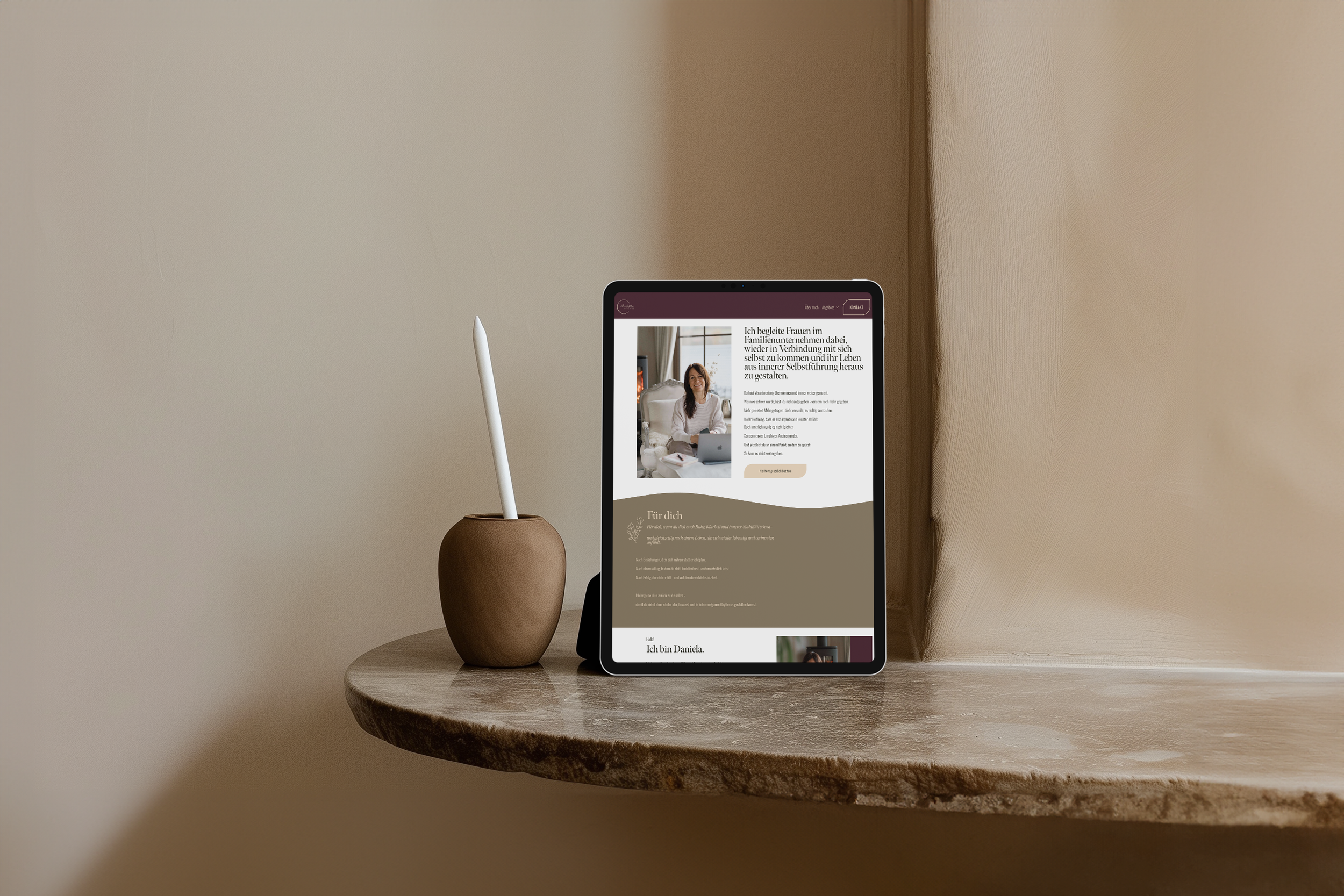 Ein Tablet auf einem Holztisch zeigt eine Webseite mit einem Bild einer Frau. Neben dem Tablet steht eine braune Vase mit einer weißen Stabblume. Die Wand im Hintergrund ist beige.