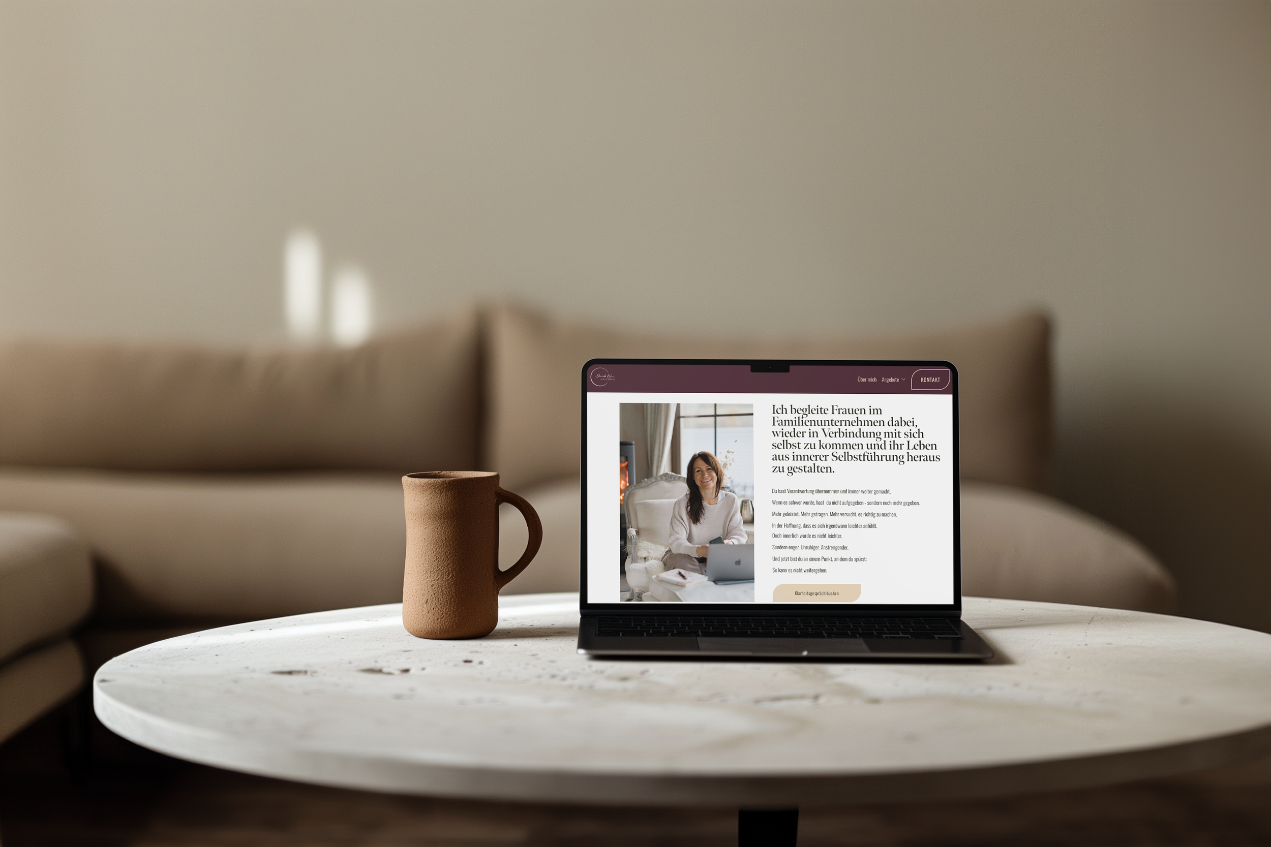 Ein Laptop auf einem weißen Tisch zeigt eine Website mit einem Bild einer lachenden Frau und deutschen Text über Selbstverbindung und Selbstführung. Neben dem Laptop steht eine braune, kerzenförmige Tasse. Im Hintergrund ist eine beige Couch und eine Wand sichtbar.