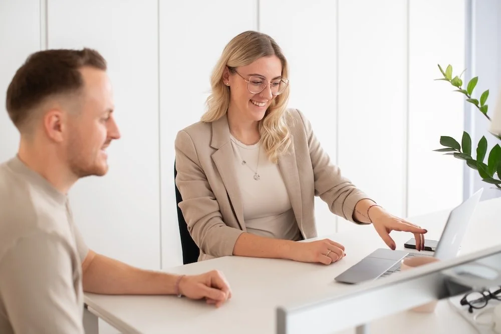 Zwei Menschen sitzen im Büro, der weibliche Assistent zeigt auf einen Laptop, beide lachen.