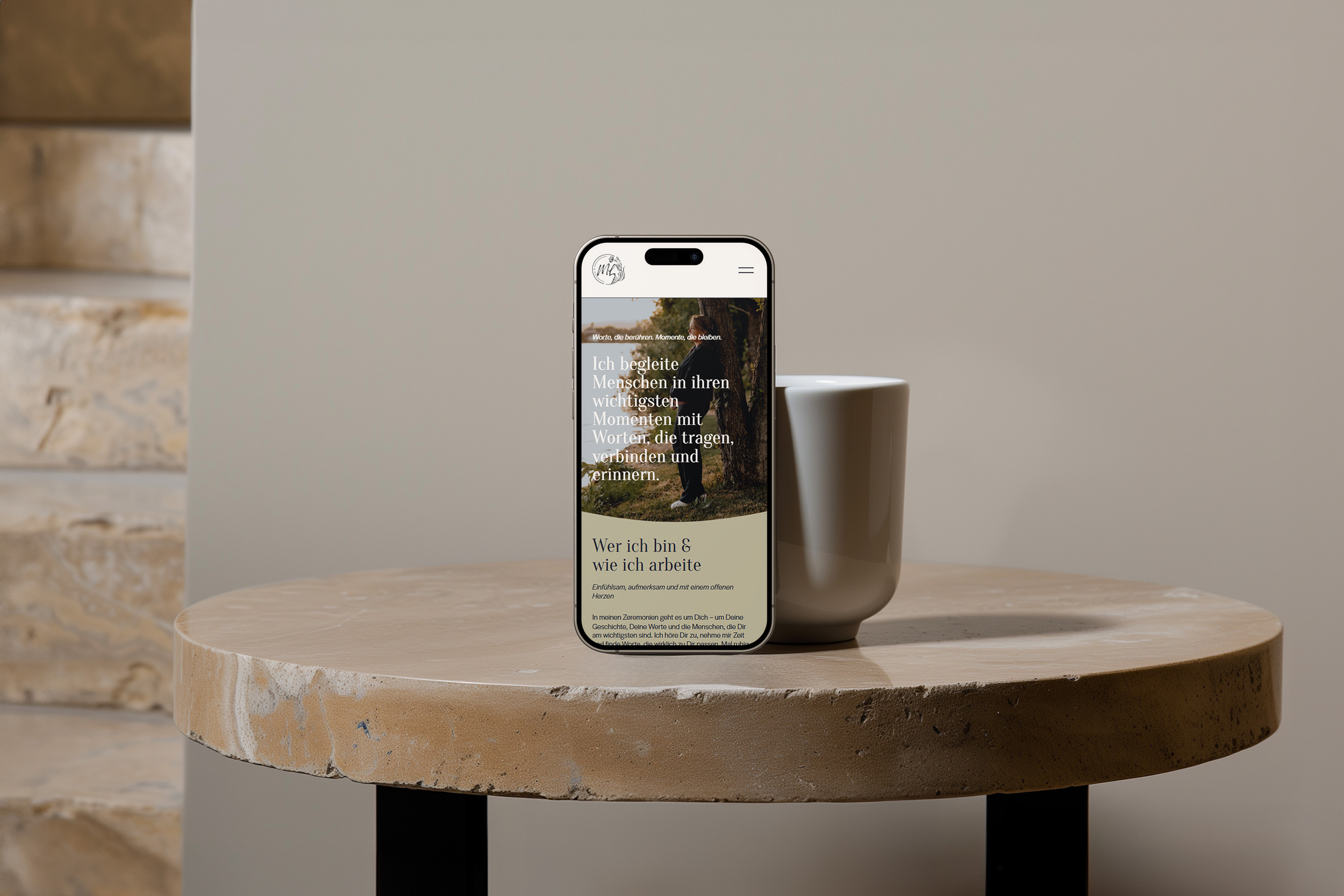 Smartphone steht auf einem runden Holztisch neben einer weißen Tasse, zeigt eine Webseite mit Text in deutscher Sprache.