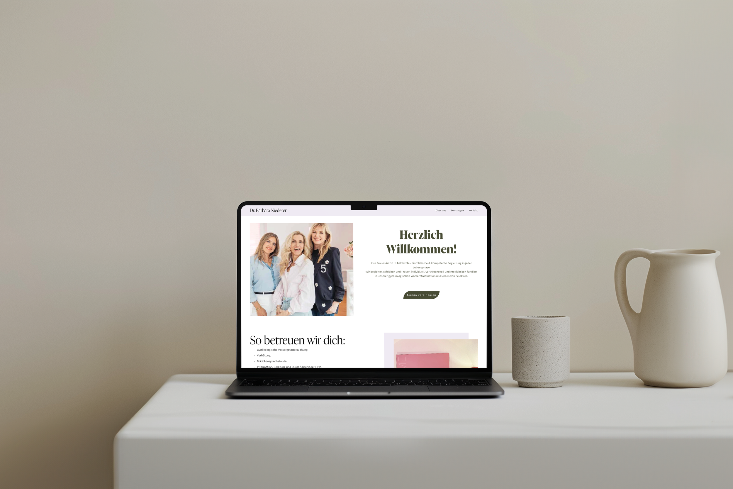 Ein offener Laptop auf einem weißen Tisch zeigt eine Webseite einer Frauenärztin mit einem Willkommensgruß und einem Gruppenfoto von drei Frauen. Daneben befinden sich eine große, runde weiße Vase und eine kleine, zylindrische Schale.