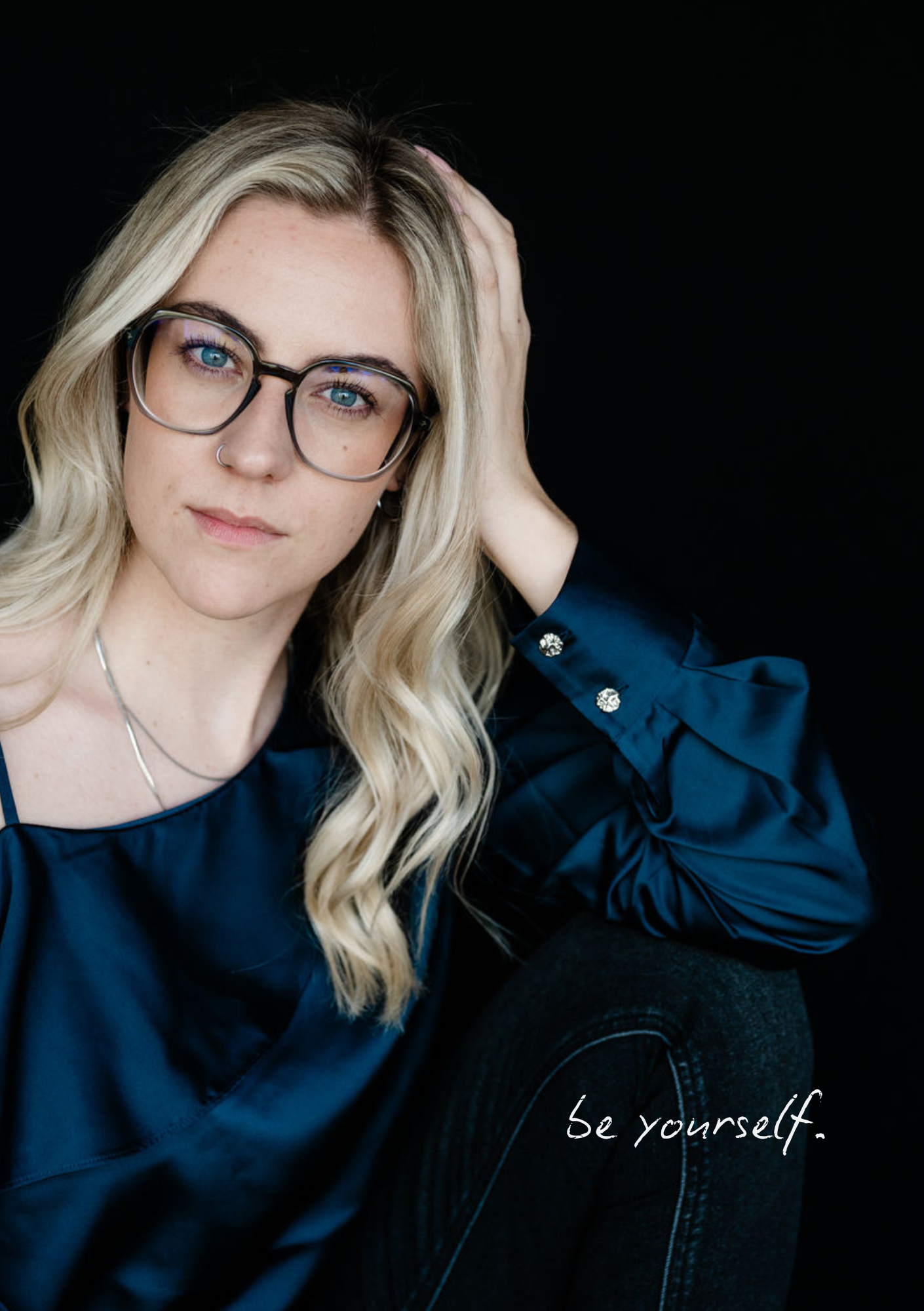 Junge Frau mit blonde Haare, Brille und einem Nasenring posiert vor schwarzem Hintergrund. Sie trägt eine dunkelblaue Bluse und schwarze Jeans. Der Text 'be yourself.' ist überlagert im unteren rechten Bereich des Bildes.