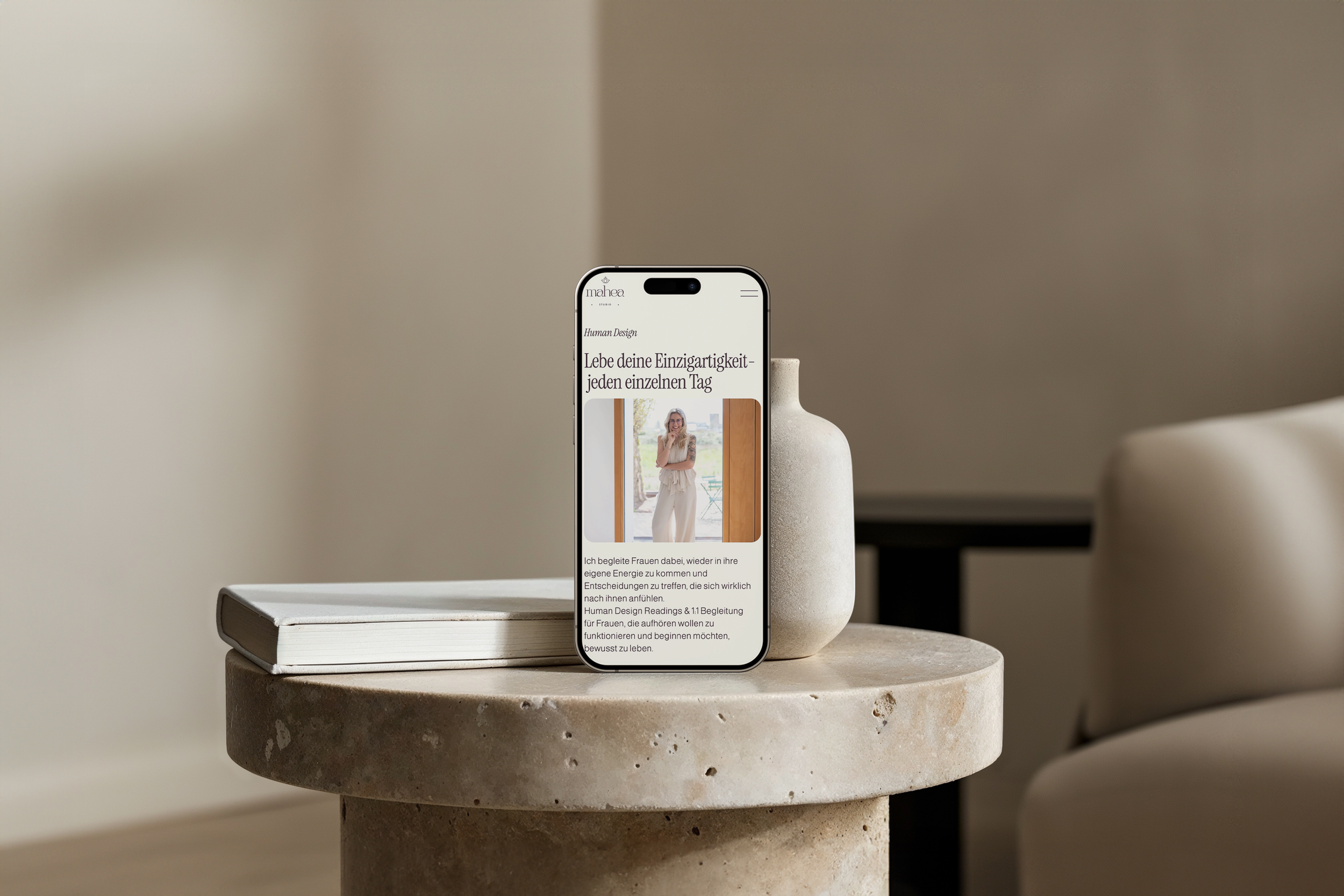 Ein Smartphone zeigt eine Website mit einem Bild einer lachenden Frau und Text in deutscher Sprache. Das Smartphone steht auf einer runden, steinernen Tischplatte, daneben befindet sich eine weiße Vase.