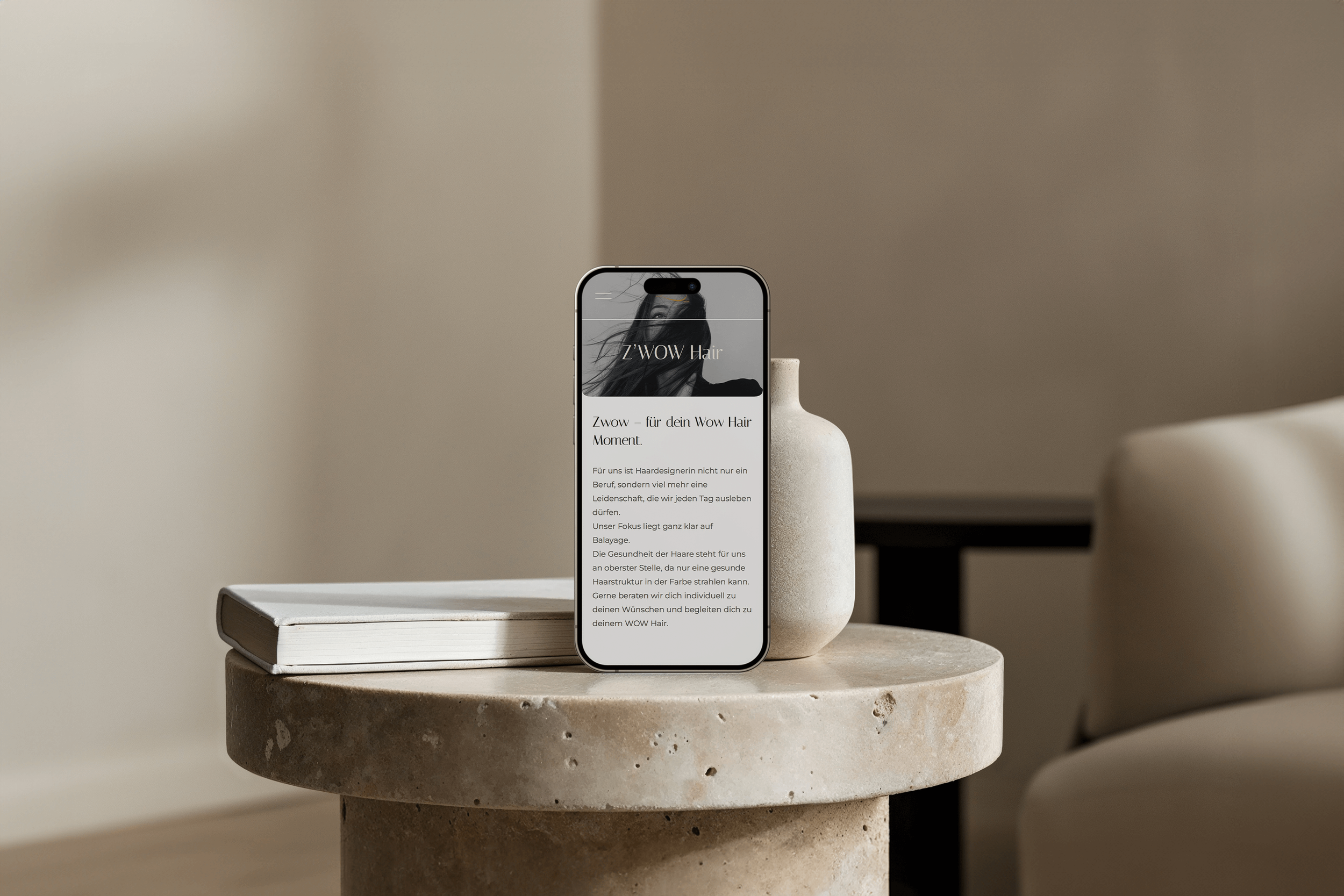 Smartphone auf einem runden Steintisch, zeigt Webseite von Z'WOW Hair, daneben ein Buch und eine weiße Vase. Im Hintergrund ein Sofa und eine dunkle Tischplatte.