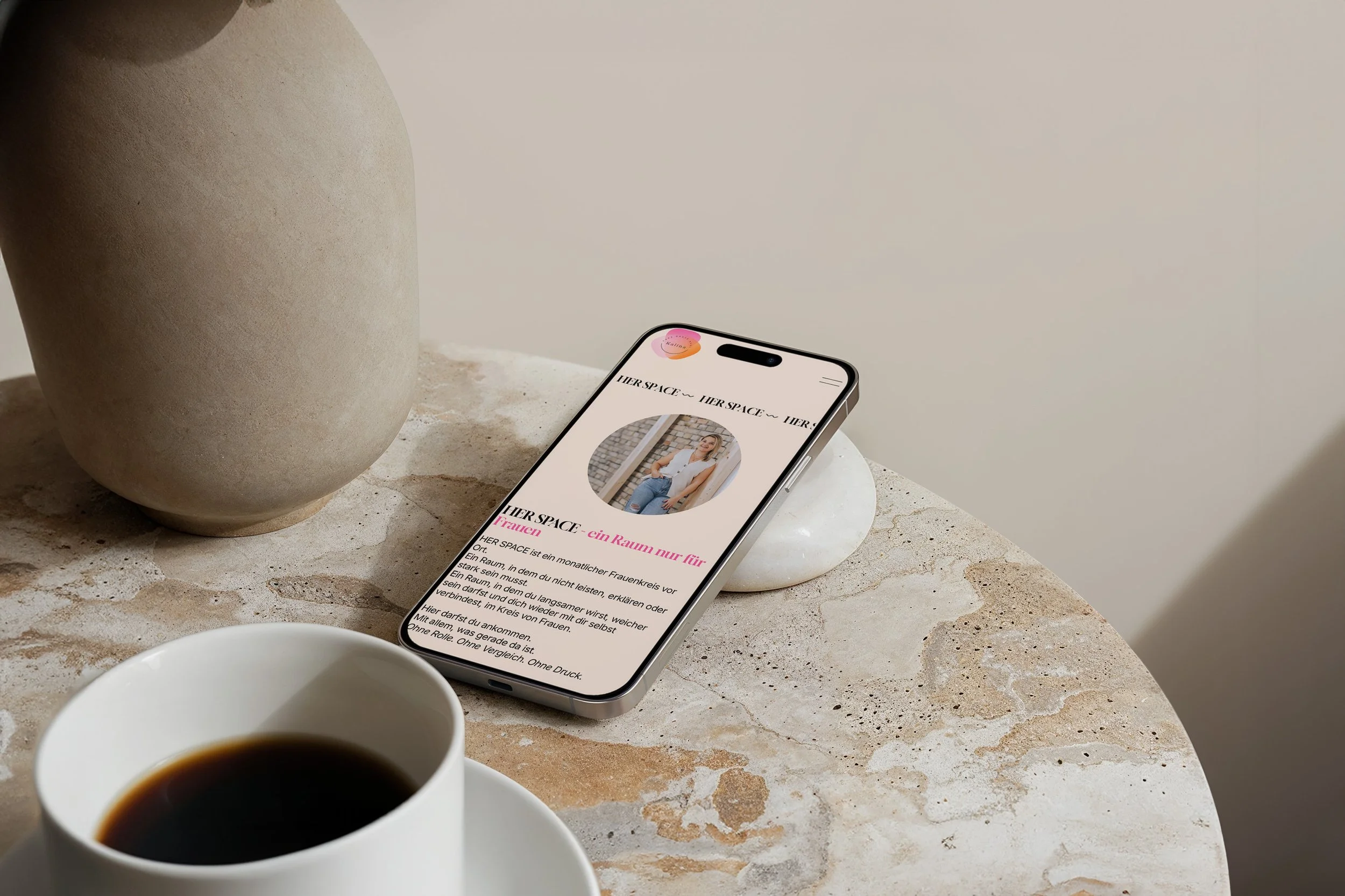 Ein Smartphone zeigt eine Webseite mit einem Bild einer lachenden Frau. Neben dem Smartphone stehen eine Kaffeetasse und eine kleine Schale auf einem Marmor-Tisch, daneben ein großer, unregelmäßig geformter Stein. Der Hintergrund ist eine schlichte helle Wand.