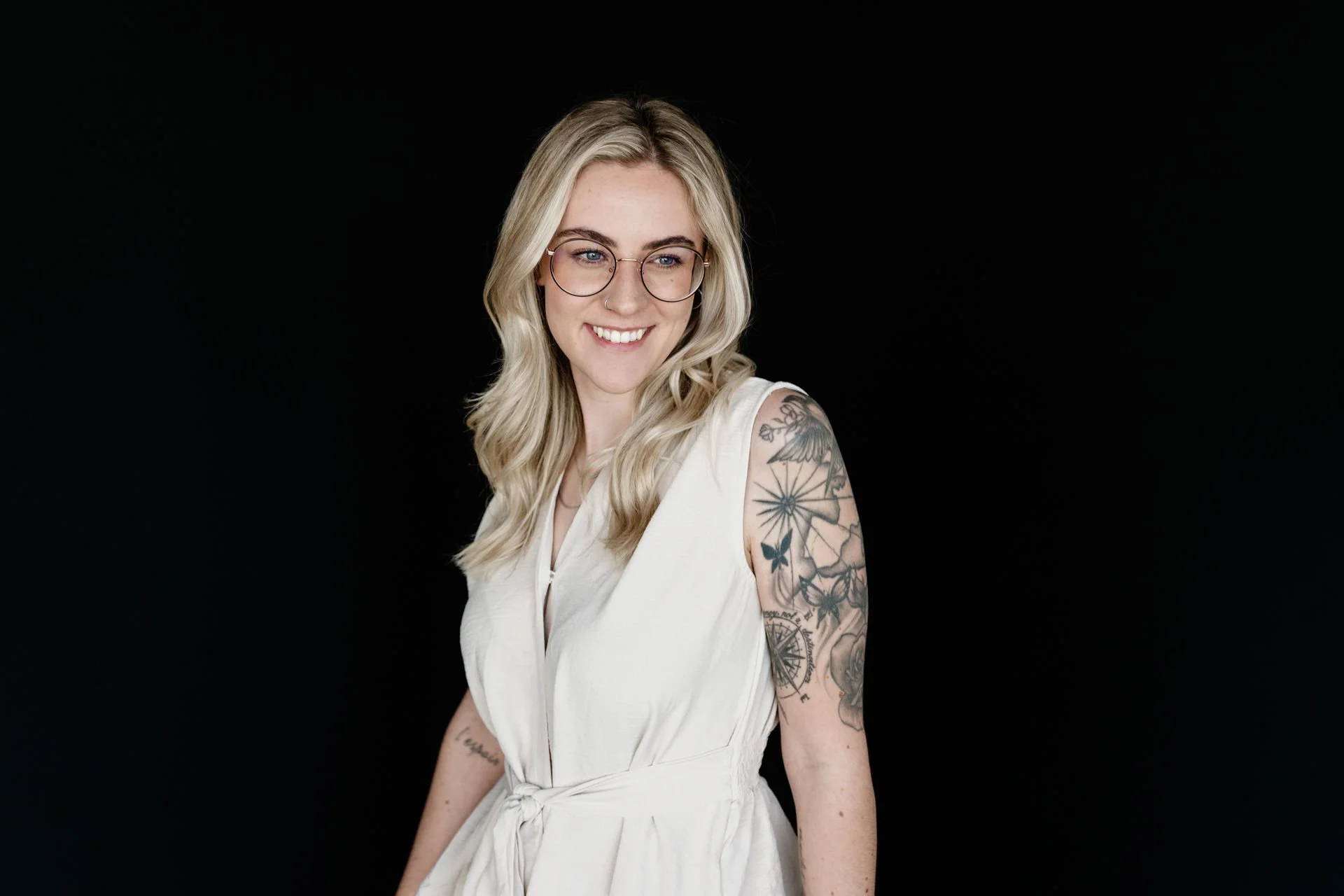 Junge Frau mit Brille und Tattoos, trägt ein weißes Kleid, lächelt vor schwarzem Hintergrund.