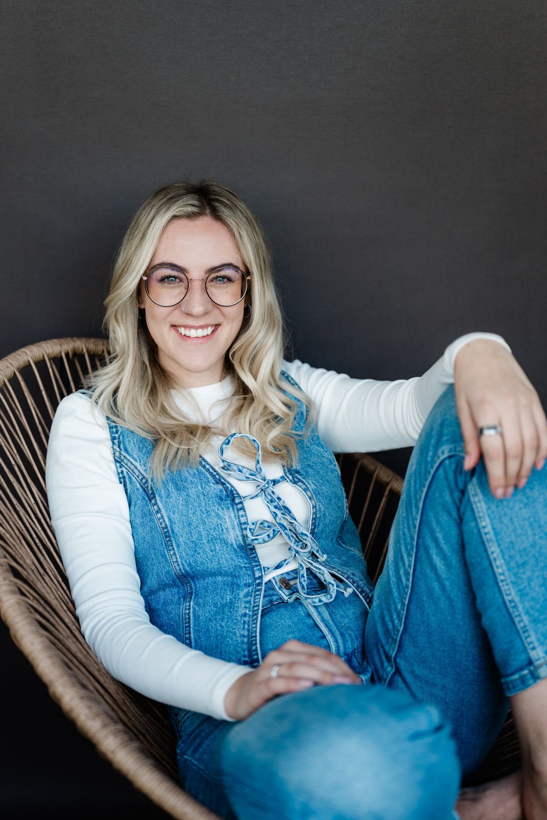 Lächelnde junge Frau mit Brille in einem weißen Langarmshirt und blauer Jeans, die in einem Korbwarenstuhl sitzt, vor einem dunklen Hintergrund.