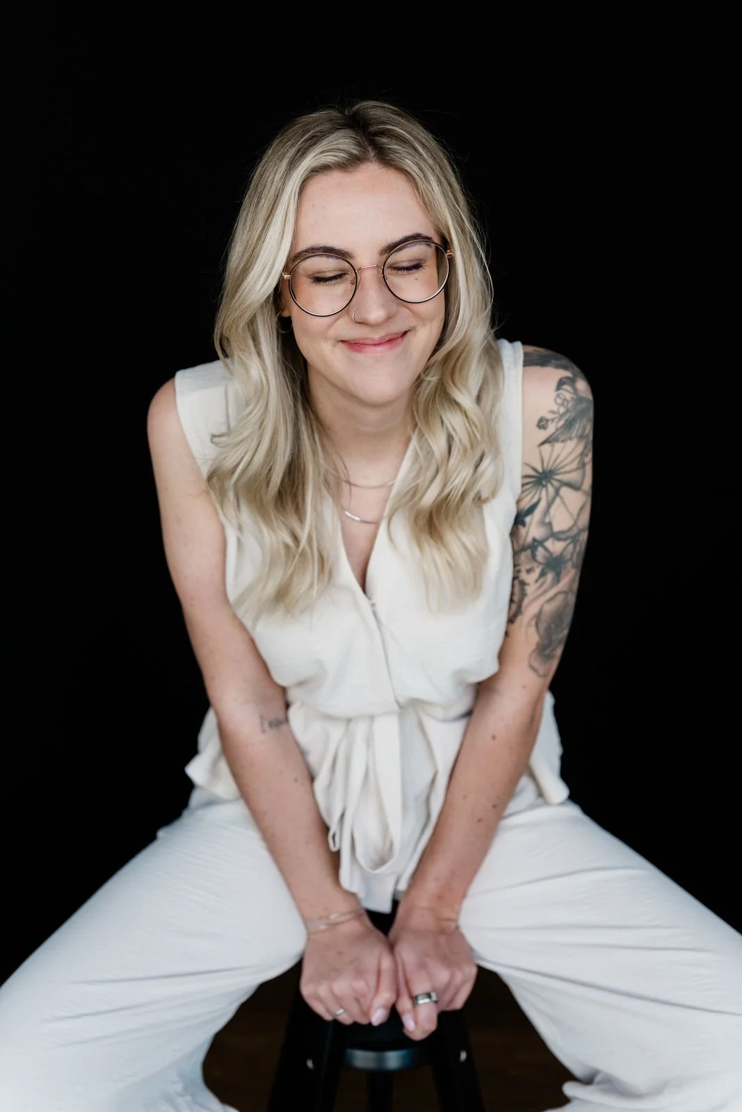 Junge Frau mit Brille, blondem gewelltem Haar, Tattoos auf dem linken Arm, lachend mit geschlossenen Augen, sitzend auf einem schwarzen Stuhl vor schwarzem Hintergrund.