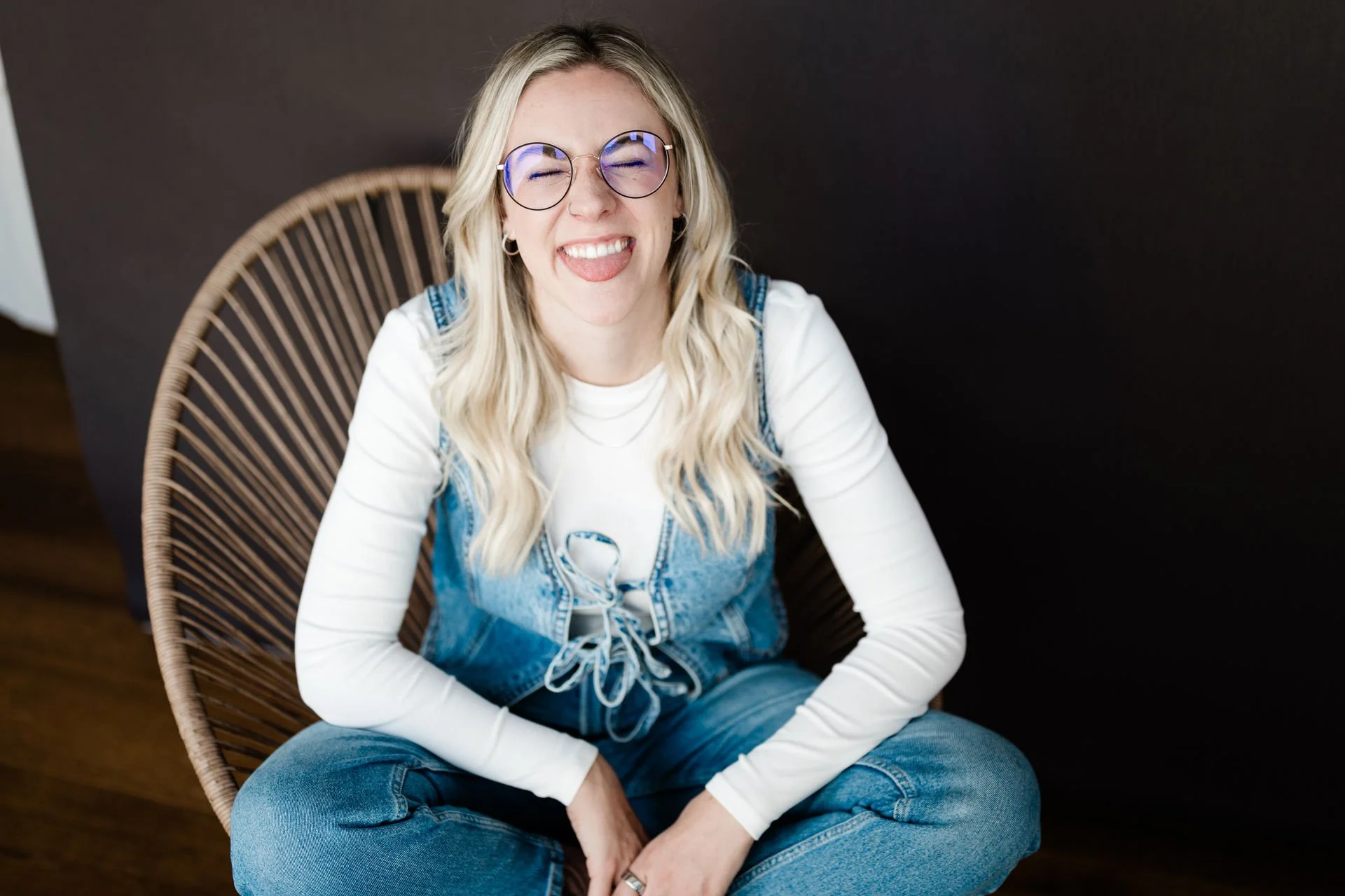 Lächelnde junge Frau mit Brille, blonde Haare, sitzt im runden Stuhl, trägt weiße Shirts, Jeansjacke, graue Jeans, positiver Ausdruck, dunkler Hintergrund.