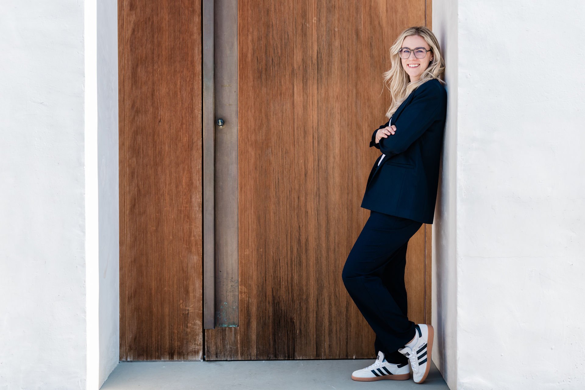 Junge Frau mit Brille, schwarze Jagdjacke, schwarze Hose, weiße Turnschuhe, lächelnd, lehnt an einer Holztür in einer weißen Wand
