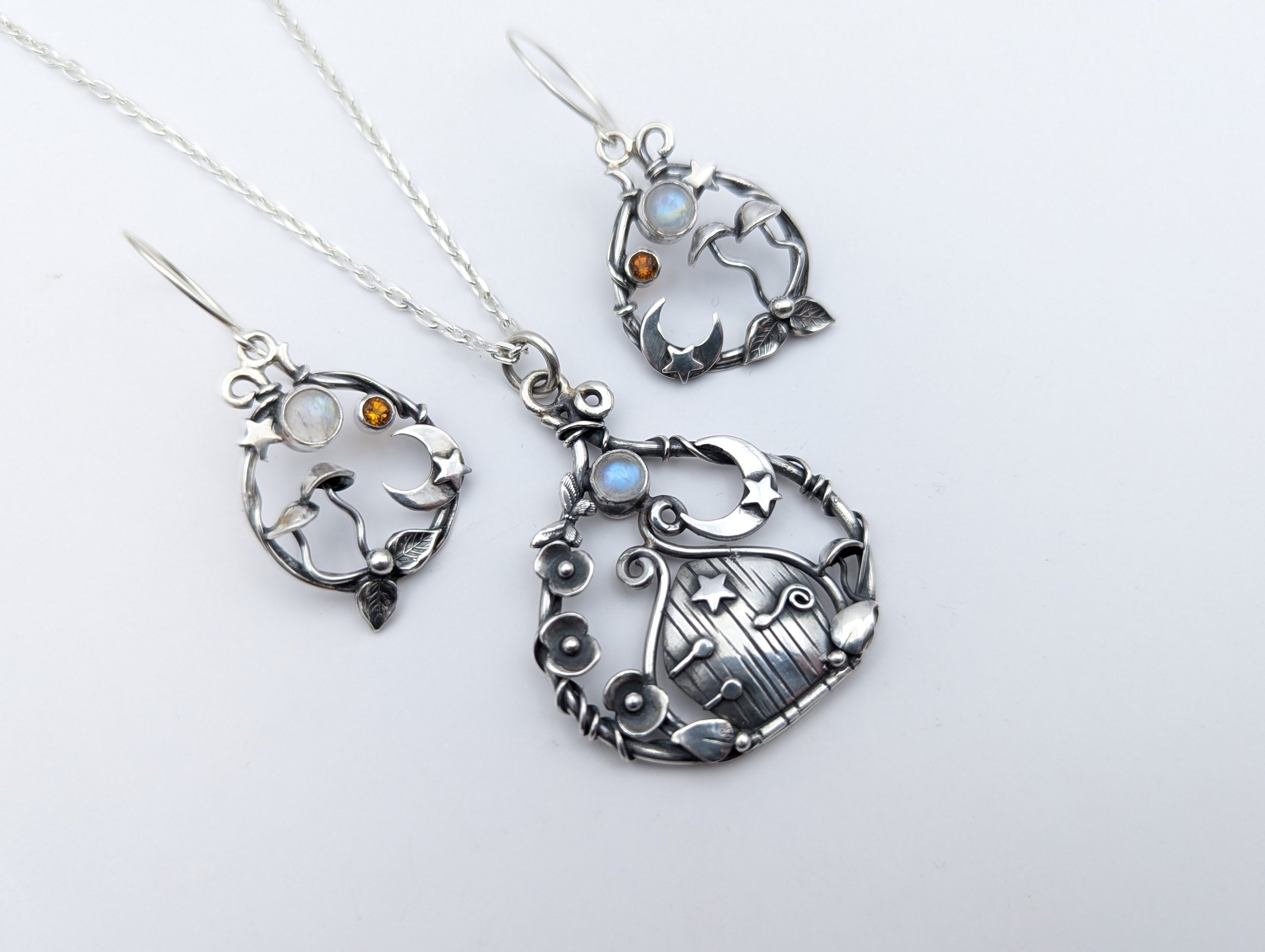 Silver jewelry set consisting of a necklace and earrings with moon and star motifs, featuring iridescent and amber-colored stones, laid against a white background.