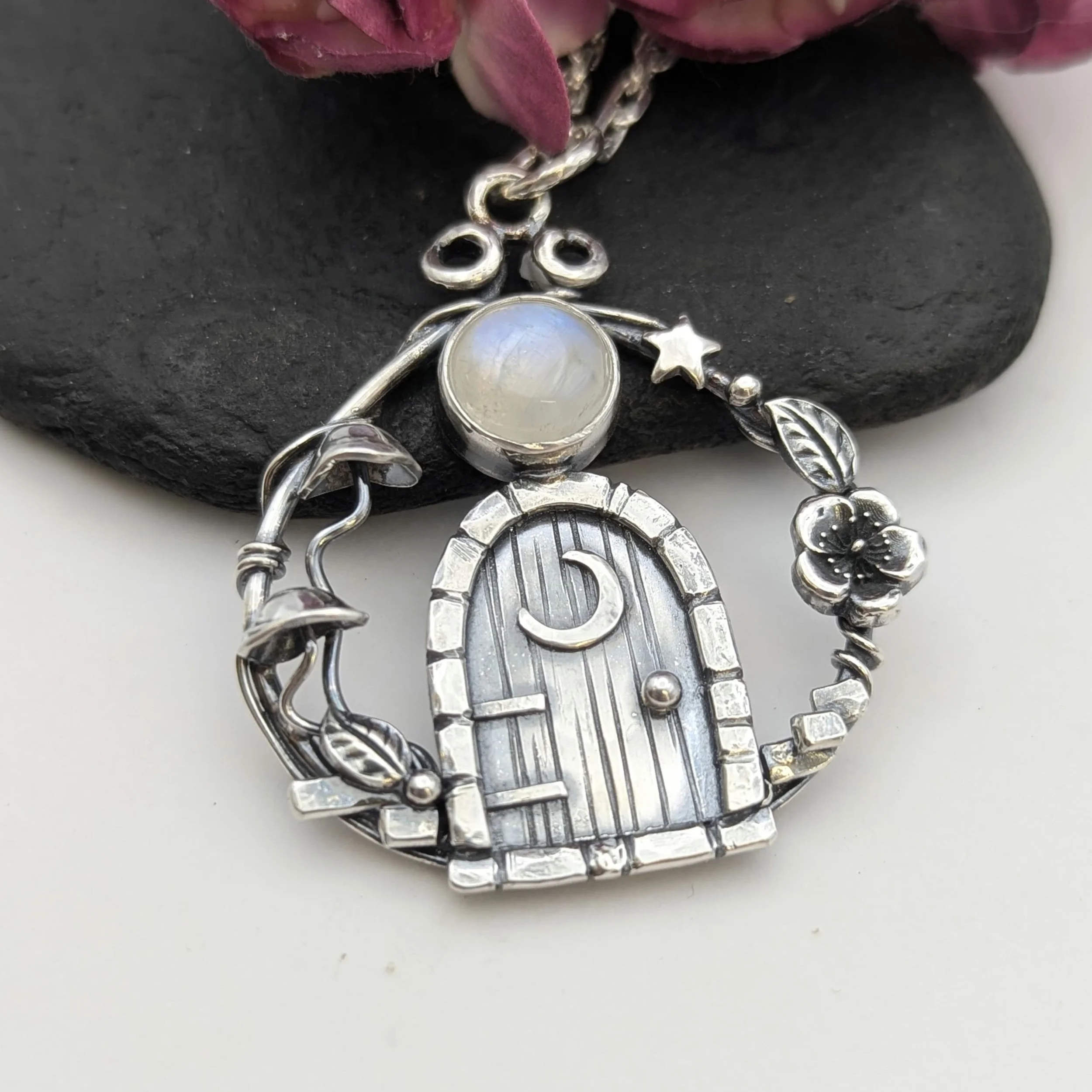 A close-up of various silver jewelry pieces including chains, rings, and charms, displayed on a black textured surface with pink flowers in the background.