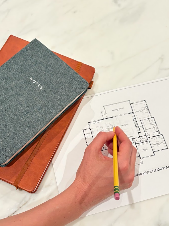 A person is marking a floor plan on a printed blueprint with a yellow pencil that has colorful stripes near the eraser. There are two notebooks, one grey with 'NOTES' on the cover and a brown one, resting on a white marble surface.