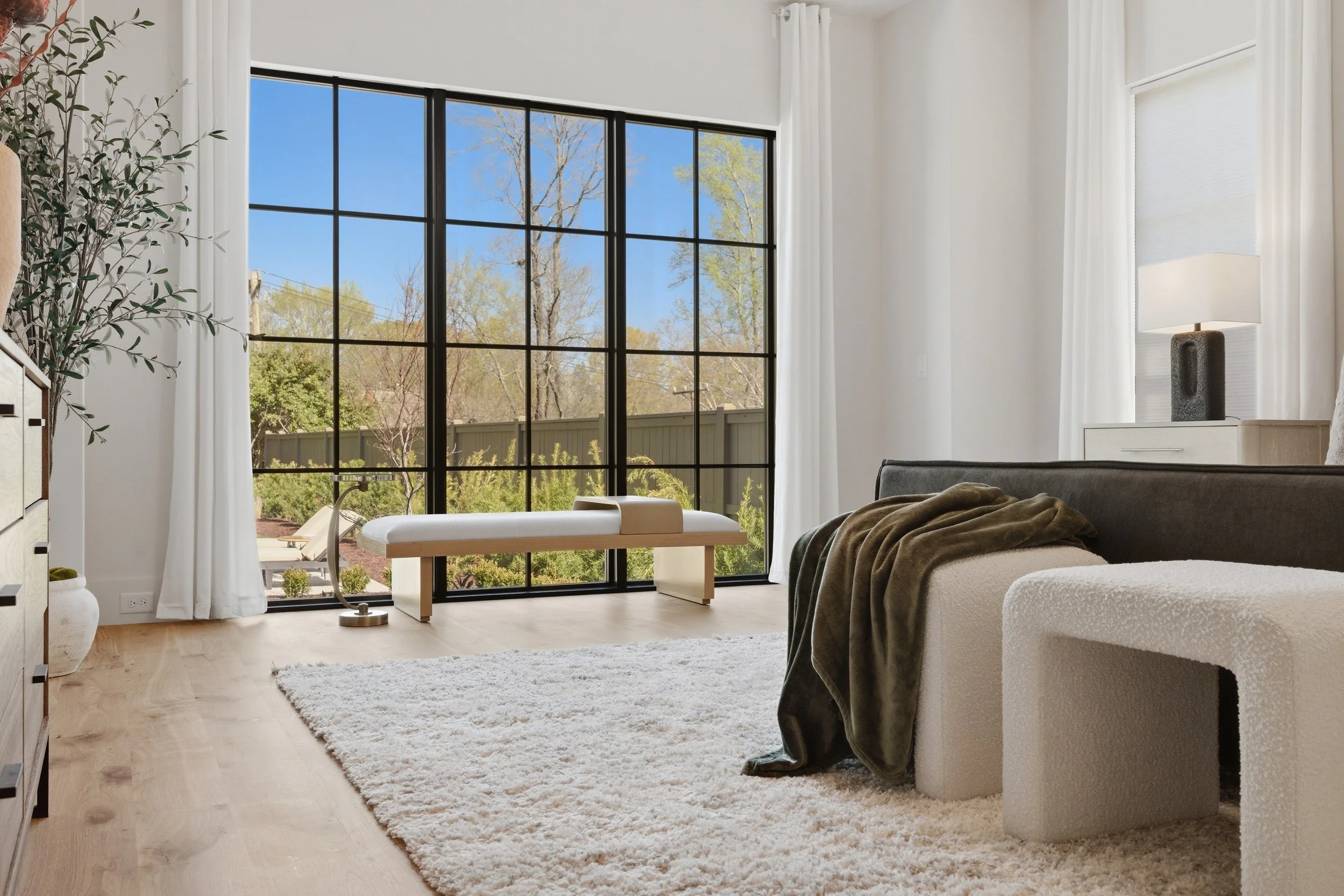 Modern bedroom with large black-framed window showing trees and a backyard, white curtains, a white dresser with a lamp, a bed with dark bedding, a white shag rug, and a potted plant.