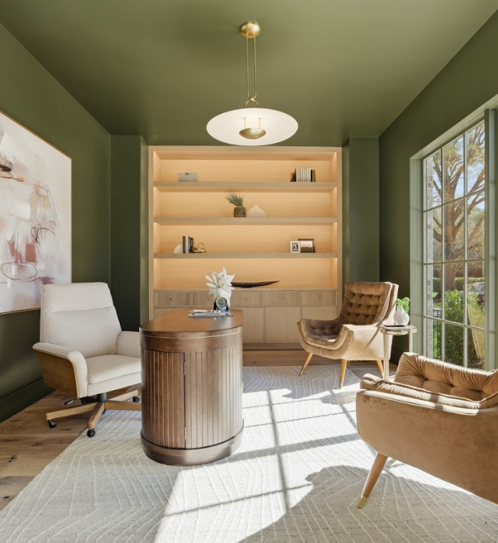 Sunlit home office with green walls, built-in light wood shelving, a curved wooden desk, and three upholstered chairs, with large window showing trees outside.