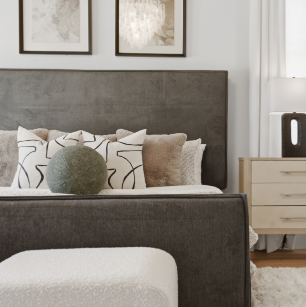 A bedroom with a gray upholstered bed, decorative pillows, and a white nightstand with a black vase, in front of white curtains.