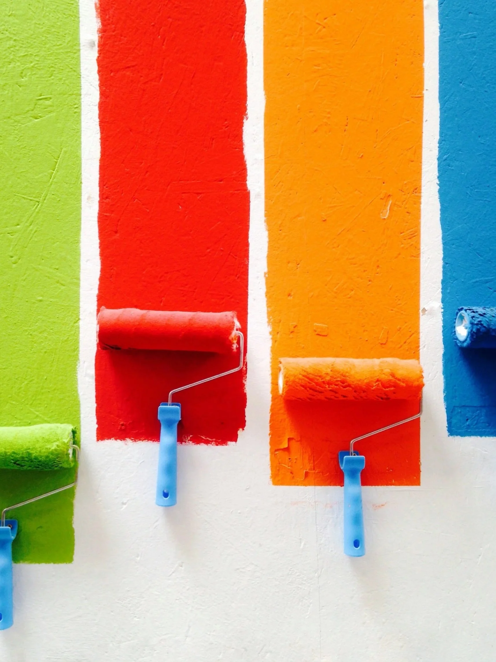 Colorful stripes painted on a wall with small paint rollers hanging from hooks.