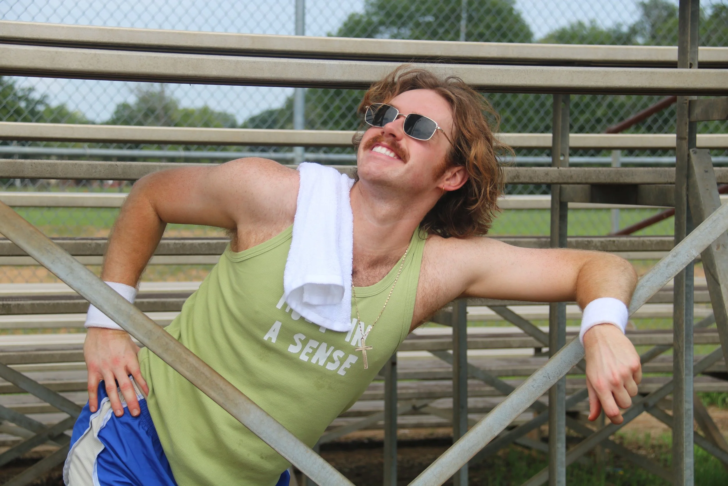 A man with long hair, sunglasses, and a mustache smiling while leaning on metal bleachers at a baseball field during daytime.
