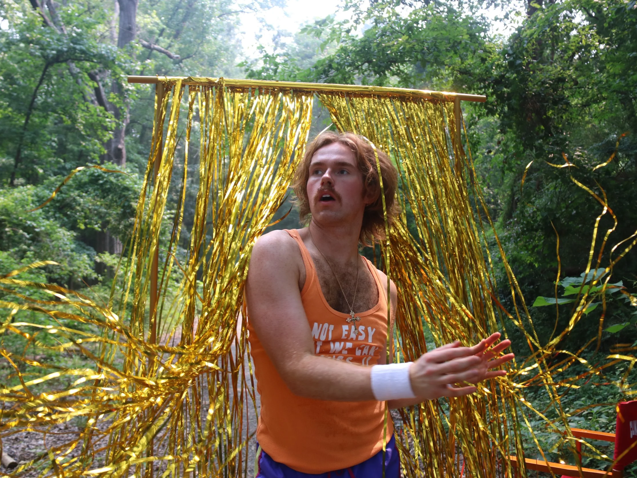 A young man with wavy hair and a mustache, wearing an orange tank top with white text and a white wristband, is standing outdoors in a wooded area surrounded by green trees. He is holding a long orange object with yellow metallic fringe hanging from a horizontal yellow pole, with his mouth open and eyes looking slightly upward.