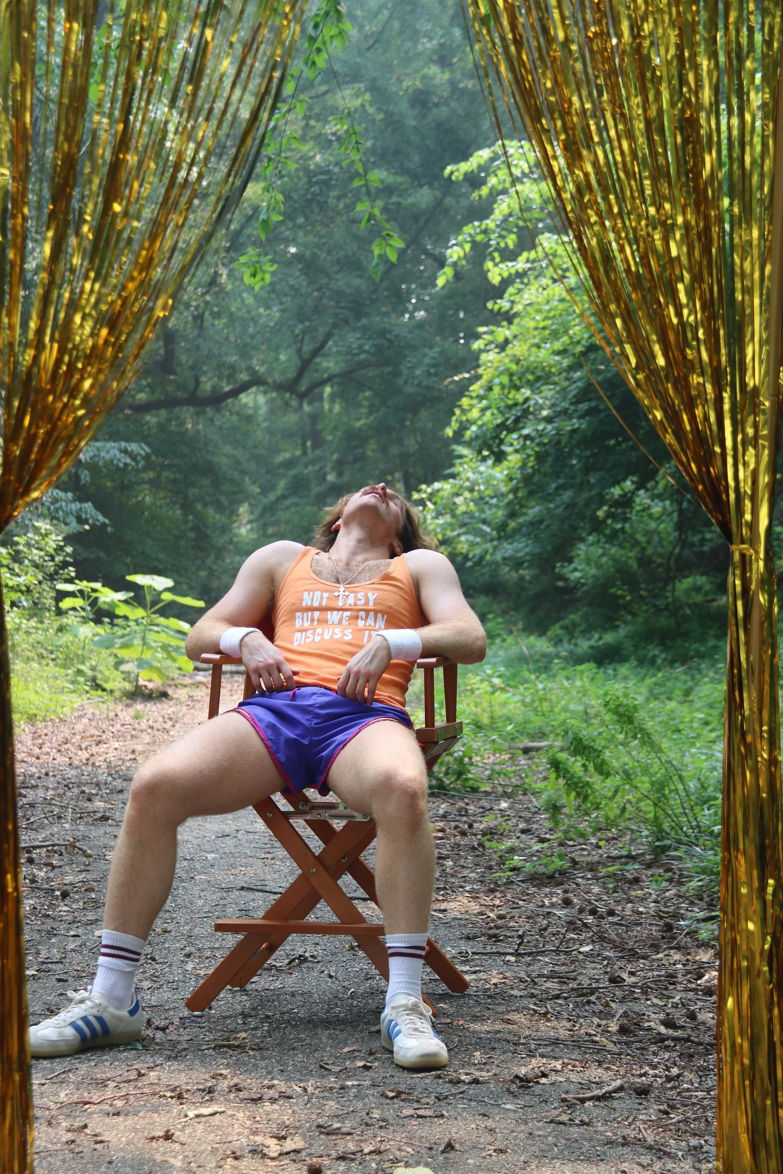 A person is sitting in a folding chair on a dirt path in a forest, with their head tilted back and eyes closed. They are wearing an orange tank top with text, blue shorts, white sneakers, and striped socks. The scene is framed by shiny gold curtains hanging on either side of the camera.