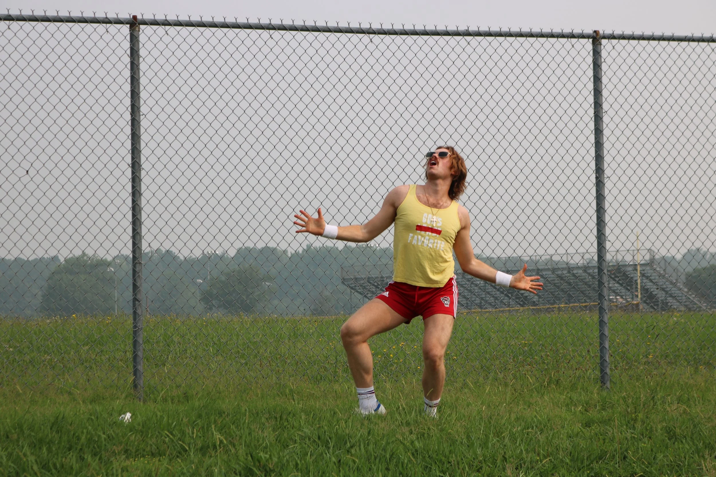 richmond based-musican clint westwood posing wearing sunglasses, a yellow tank top, and red shorts, standing in front of a chain-link fence on a grassy field, with his arms outstretched and mouth open.
