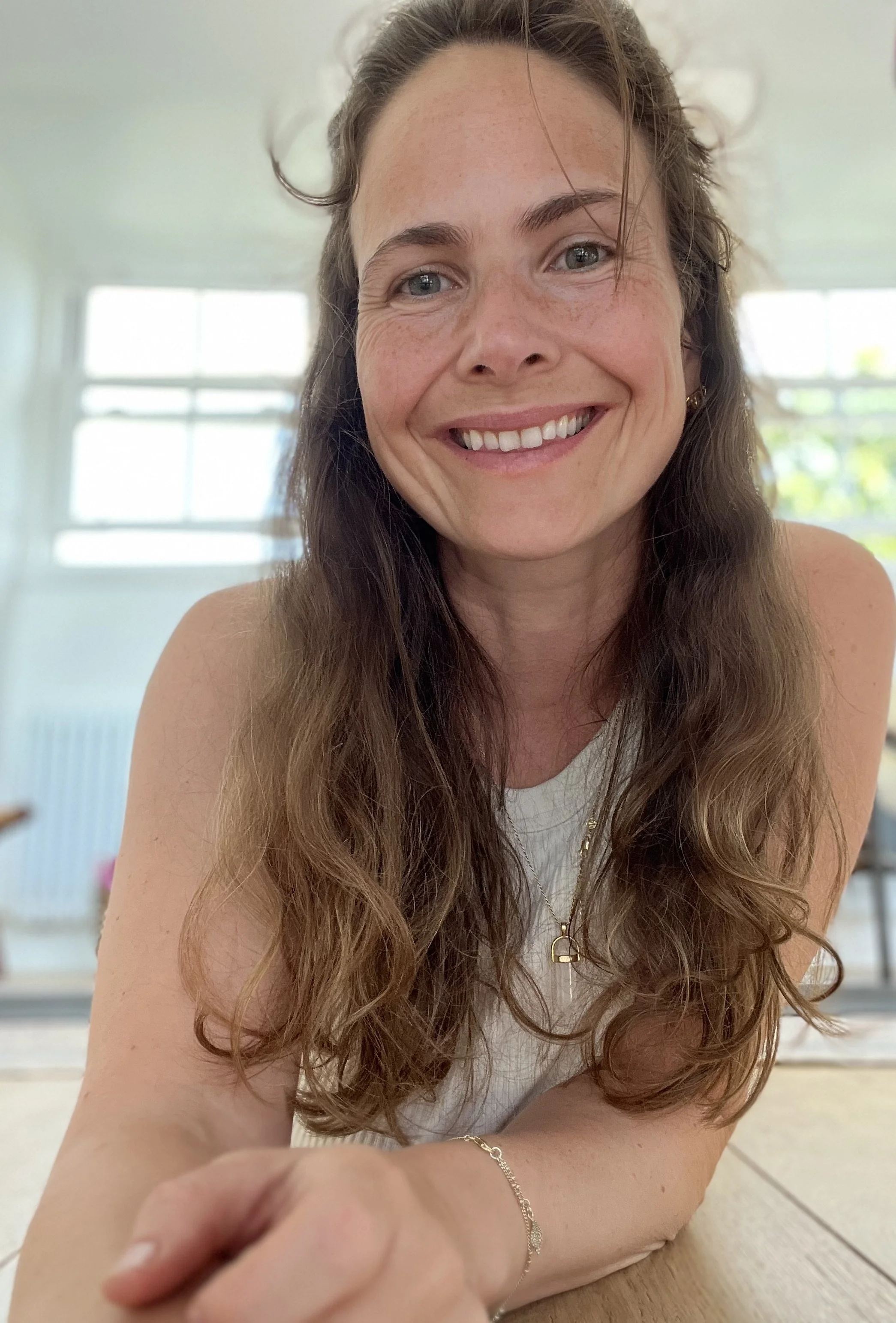 Close-up of a smiling woman with long, wavy brown hair, blue eyes, and fair skin, sitting indoors with large windows in the background.