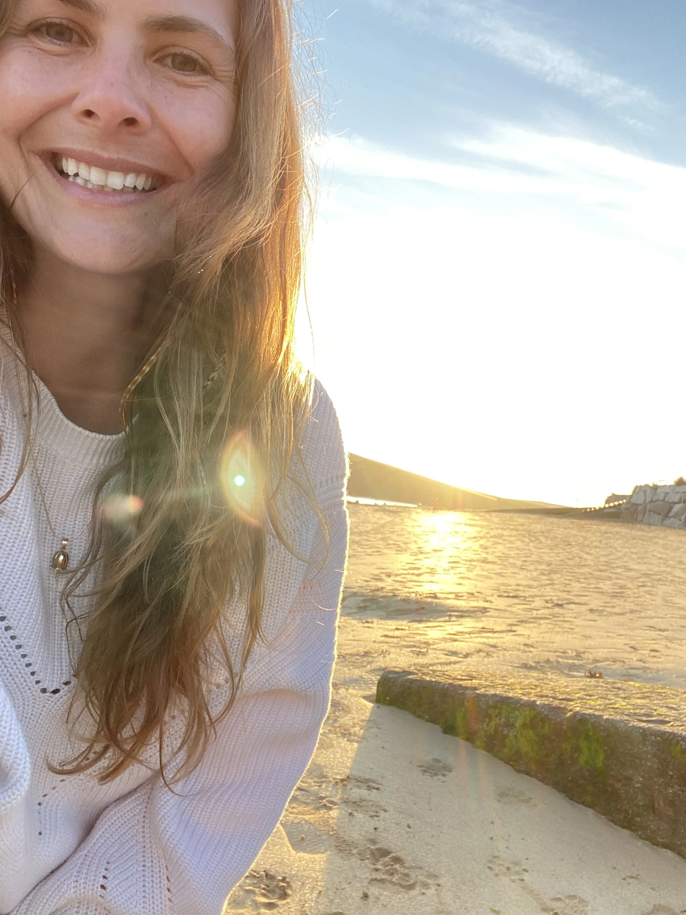 Close-up of a woman smiling on a beach during sunset, with sandy beach and rocks in the background.