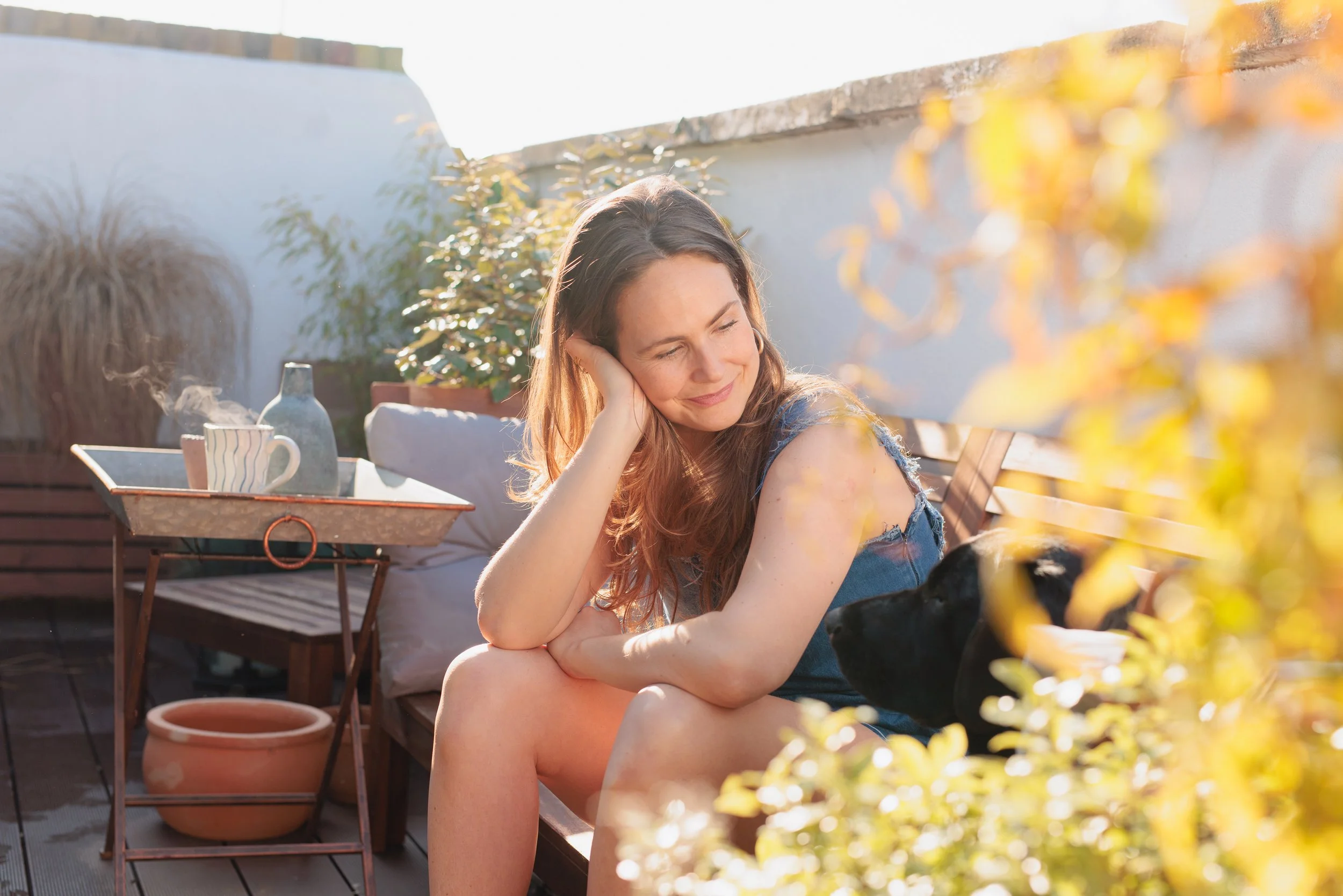 A woman with long, wavy brown hair relaxing outdoors, smiling gently with her eyes closed, leaning her head on her hand. She is sitting near a flower bed with yellow leaves and is dressed in a sleeveless blue top. The setting appears to be a sunny patio with a wooden table holding a mug and a glass pitcher, with a white wall and greenery in the background.