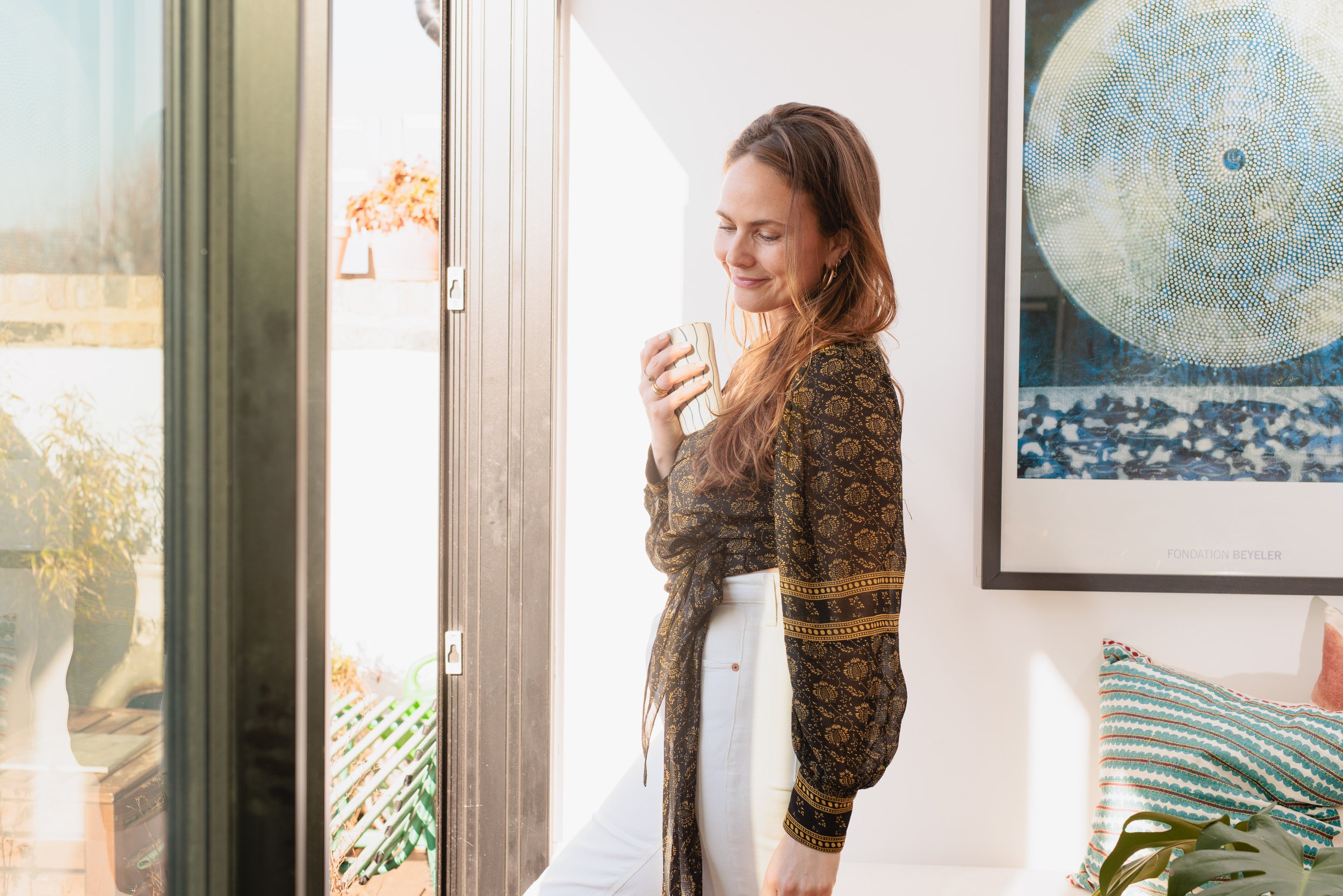 A woman with long brown hair smiling and holding a striped mug stands near a doorway with sunlight streaming in. She is wearing a black and gold patterned jacket with white pants, inside a room decorated with artwork and pillows.