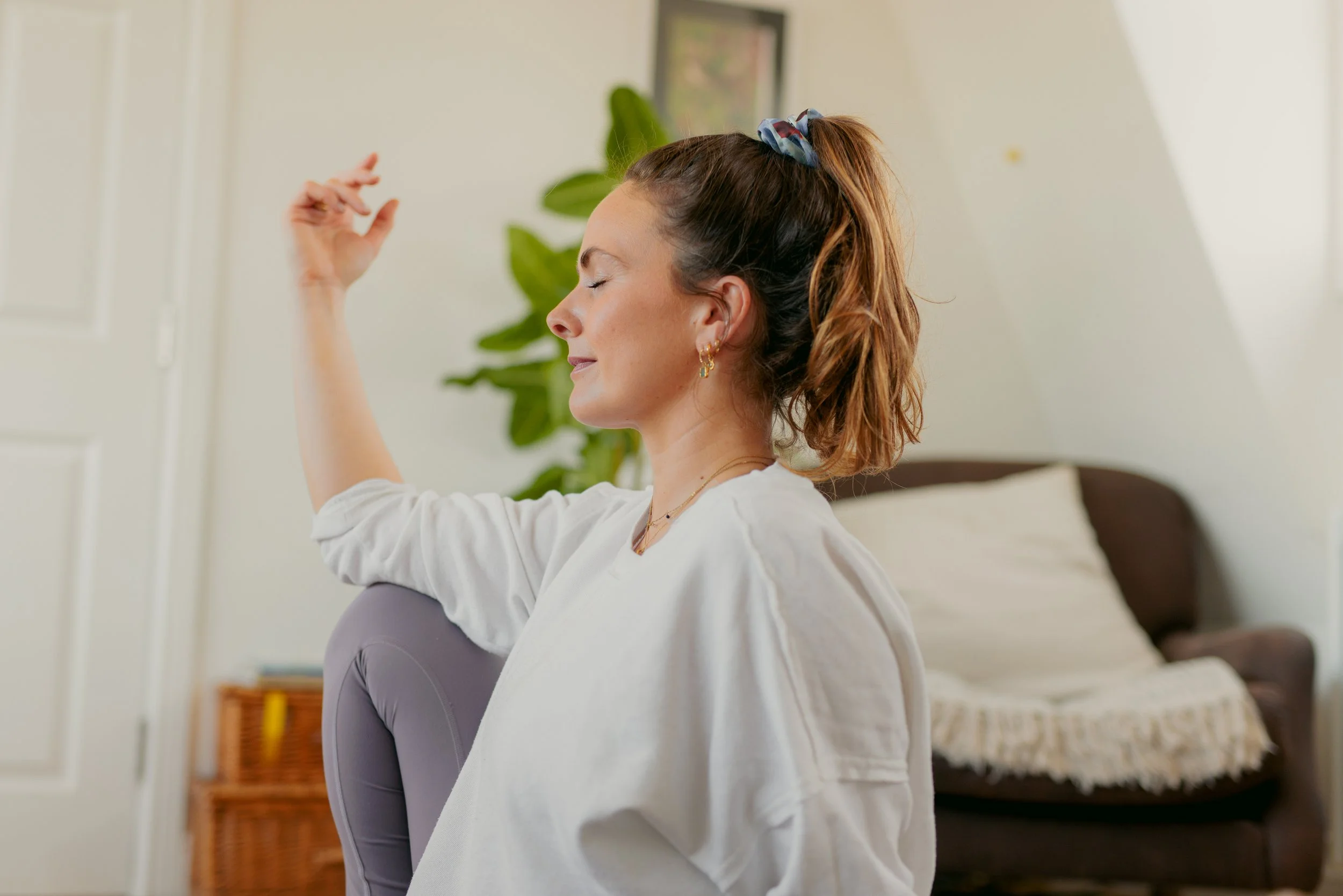 A woman with brown hair tied back with a scrunchie, sitting with eyes closed and a peaceful expression, in a cozy room with a bed, a large leafy plant, and a framed picture on the wall.