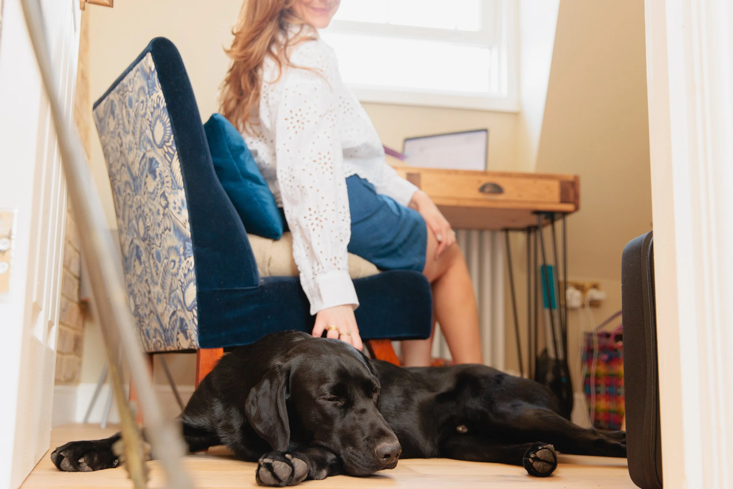 Two black Labrador puppies lie on the floor with their eyes closed, sleeping near a woman who is sitting on a chair. The woman, with red hair, is smiling and wearing a white eyelet blouse and a blue skirt, in a well-lit room with a window and a wooden desk in the background.