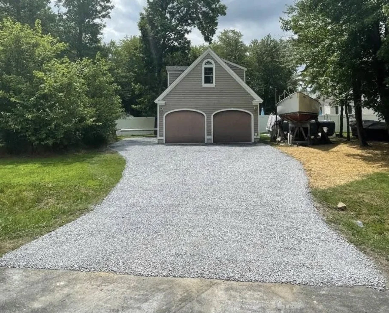 A gravel driveway leading to a two-car garage with tan doors, a small upper window, and surrounded by green trees on both sides.