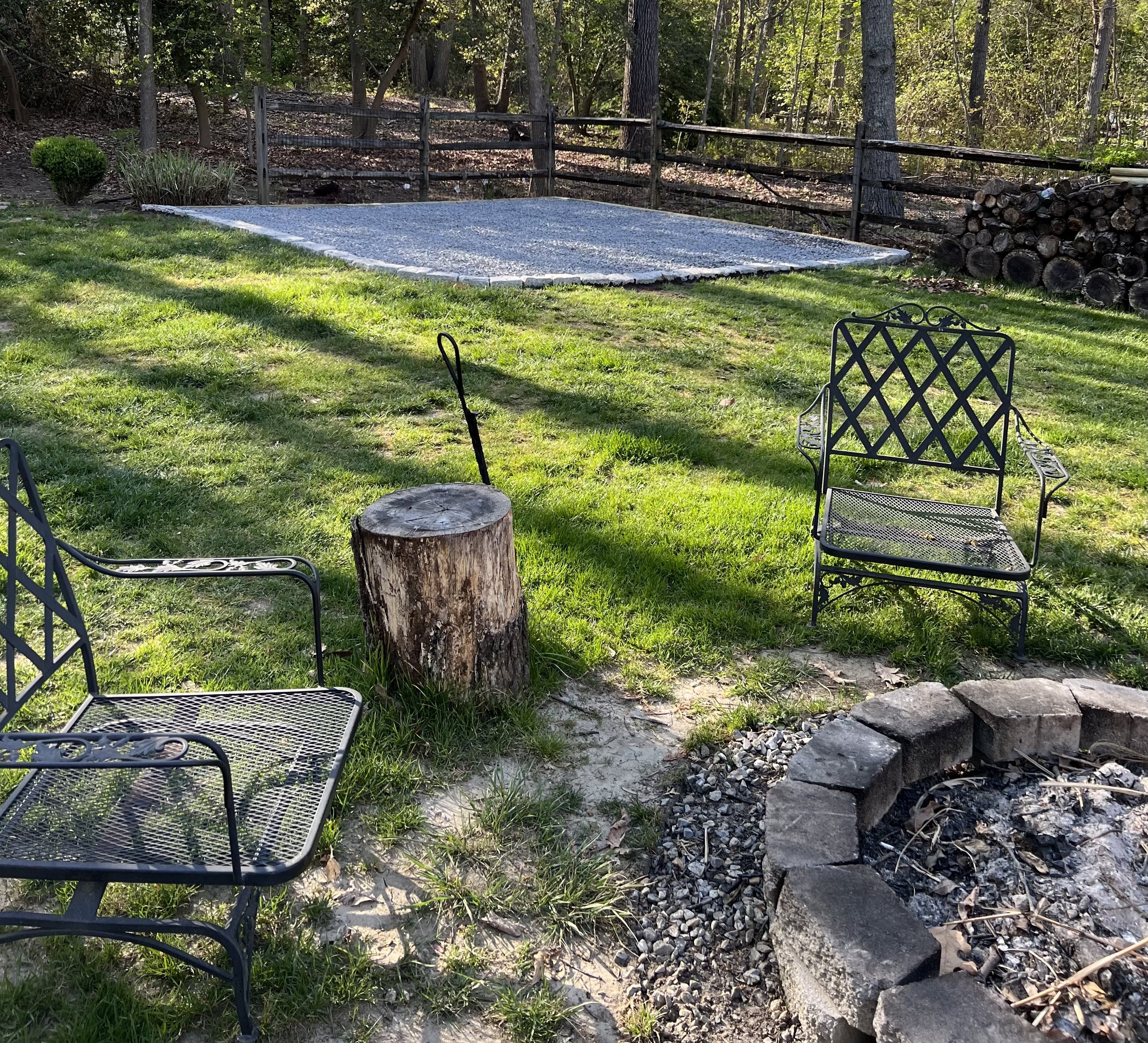Yard with a fire pit area, surrounded by lawn, trees, and a wooden fence, with firewood stacked on the side.