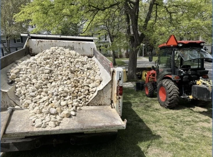 A trailer filled with white and light-colored rocks parked on grass next to a black and red tractor with a yellow attachment in a residential yard, with trees and houses in the background.