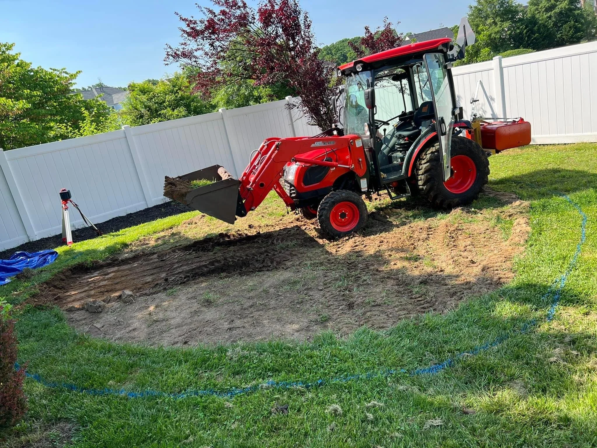 A red tractor with a front loader attachment digging into the ground in a backyard enclosed by a white fence. There are green trees and bushes in the background, and the sky is clear and blue.