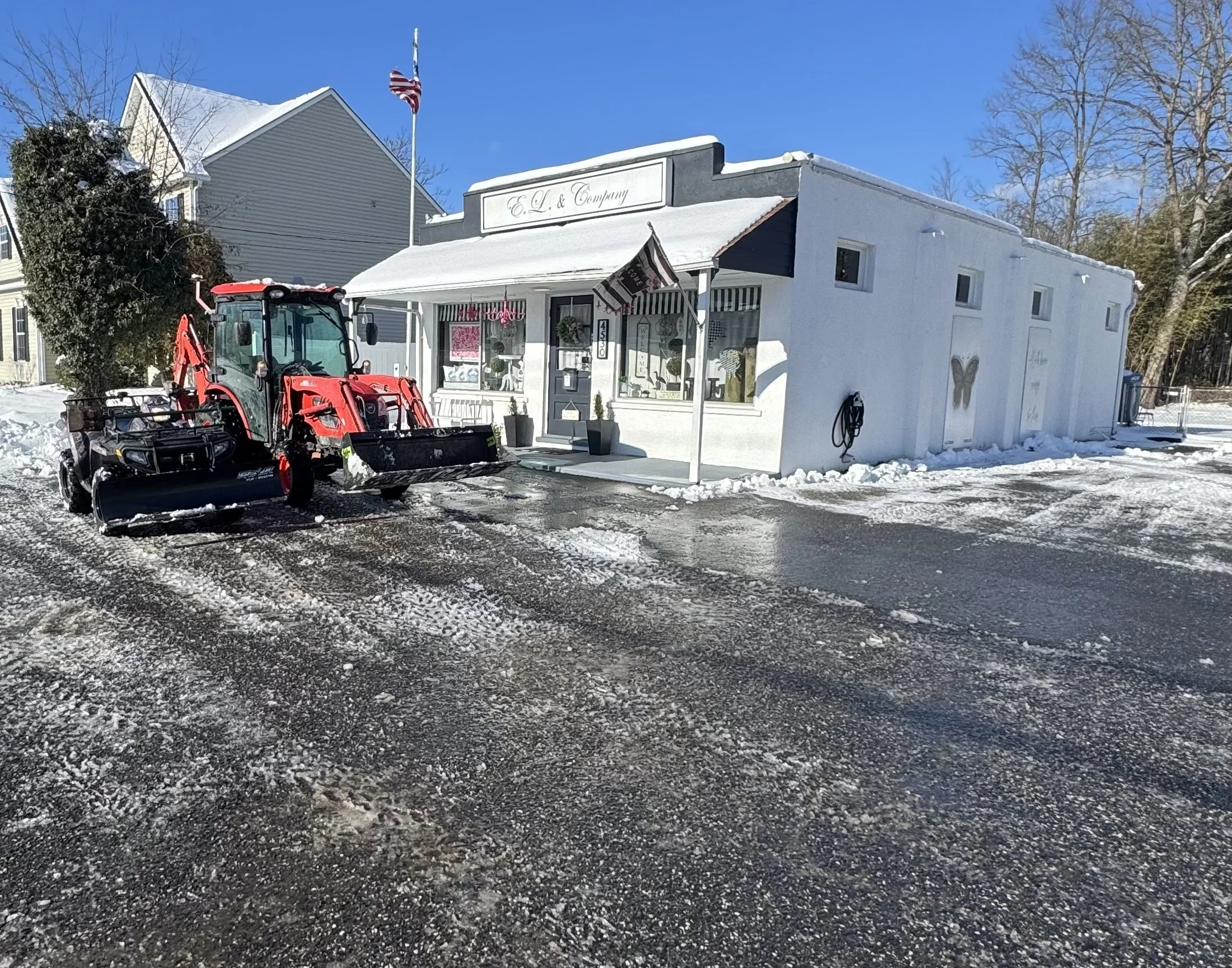 A small white building with a striped awning and a sign that reads 'E.L. & Company' is shown. A red snow removal tractor is parked in front of the building on a snowy street. An American flag is flying on a flagpole near the building. Bare trees and a bright blue sky are in the background.
