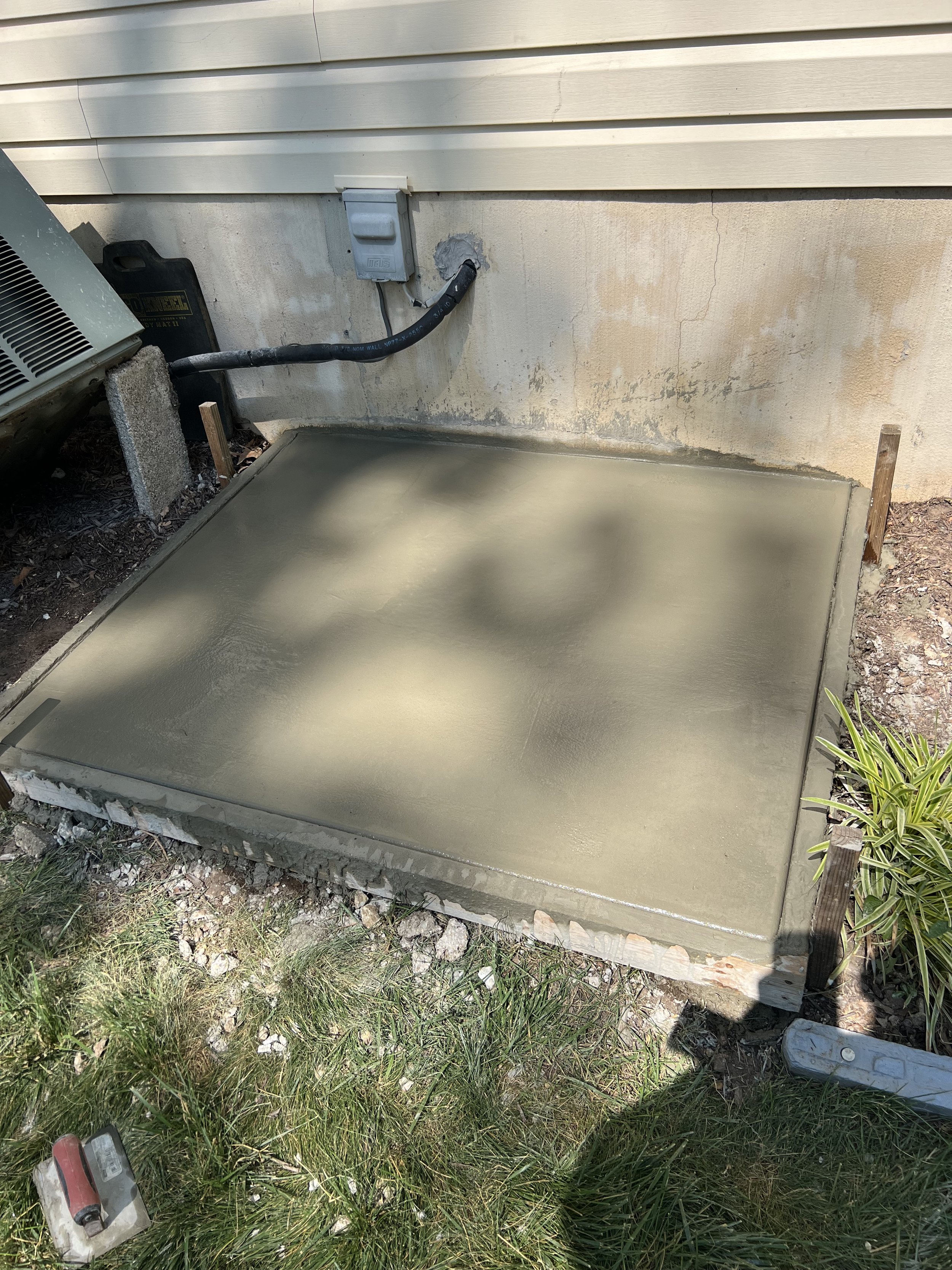 Freshly poured concrete slab with tools and construction materials nearby, outdoors next to a house wall with beige siding before finishing touches.